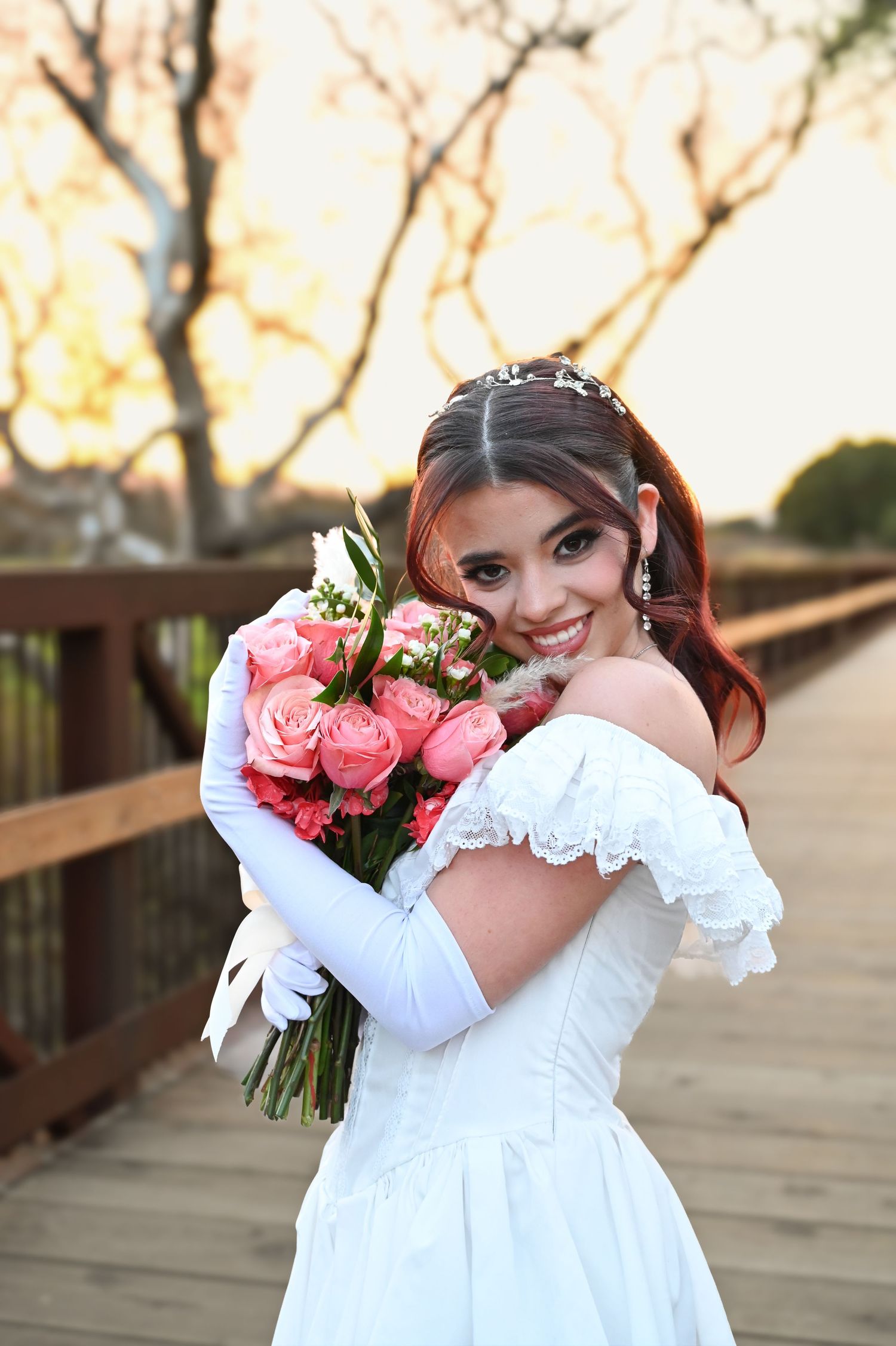 Bride in white off-shoulder gown poses with pink bouquet on wooden bridge at sunset.