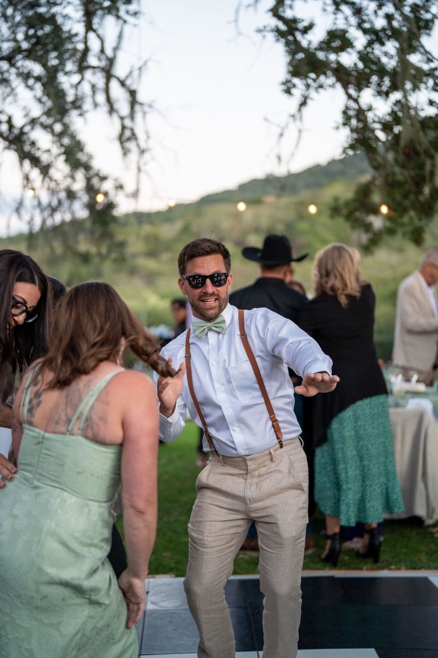 Wedding guests in mint green bridesmaid dresses and groomsmen in suspenders dance at an outdoor reception.