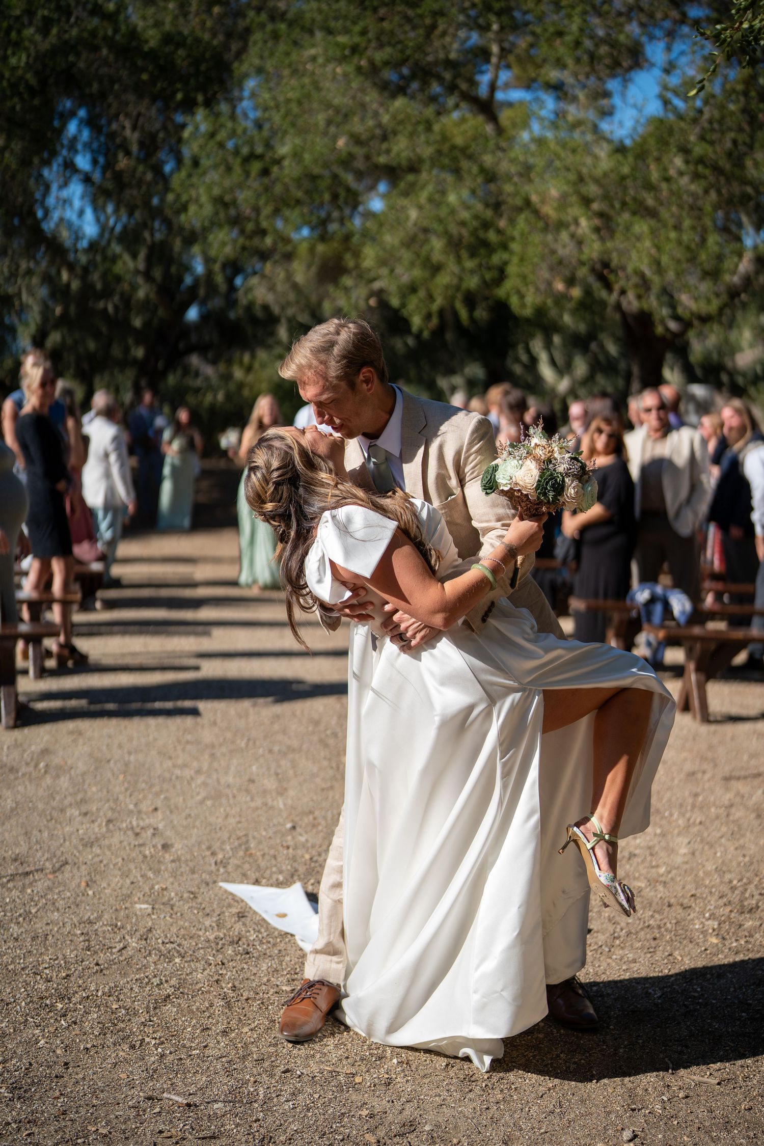 A romantic dip pose during an outdoor wedding celebration with guests gathered in the background on a sunny day.