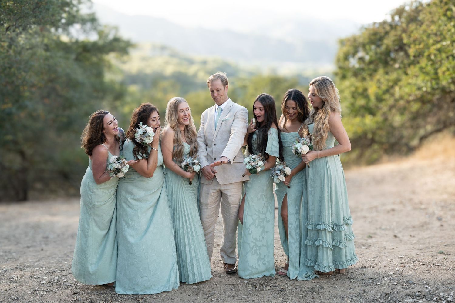 A wedding party in mint green dresses poses together on a sunny hillside path during golden hour.