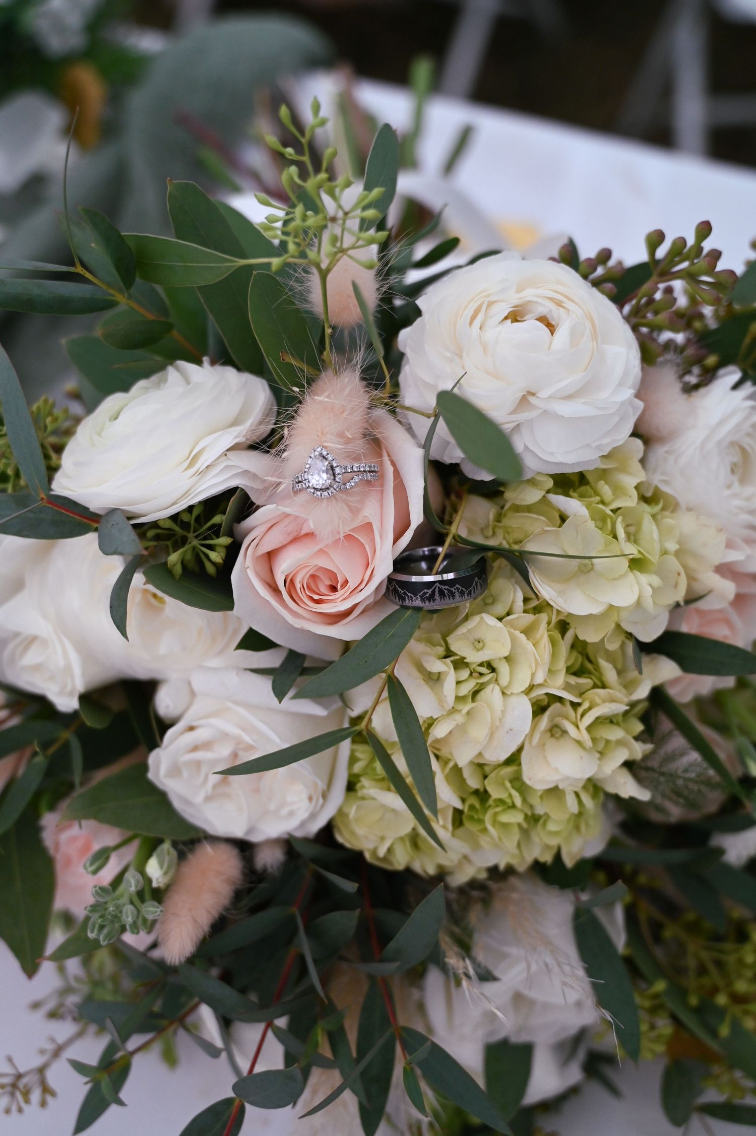 Close-up of elegant wedding rings nestled among blush pink roses, white flowers, and eucalyptus greenery in a bridal bouquet.