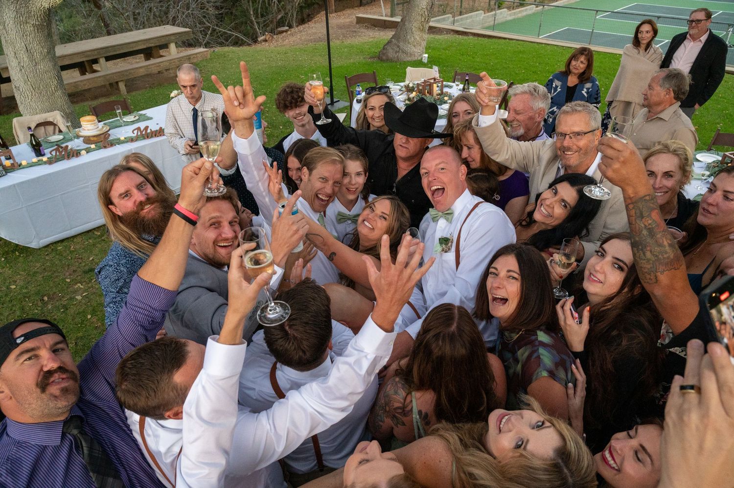 A lively group of wedding guests raise their glasses in celebration during an outdoor reception party.