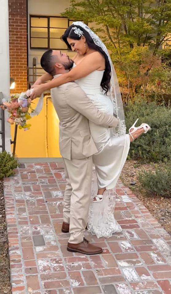 A romantic wedding moment captured on brick pavement as a couple embraces in front of a yellow door and autumn foliage.