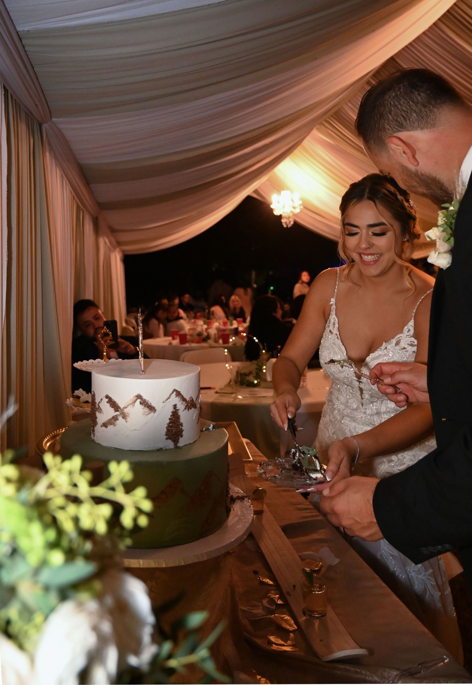 A couple cuts their elegant white wedding cake together during an evening reception under draped fabric decor.
