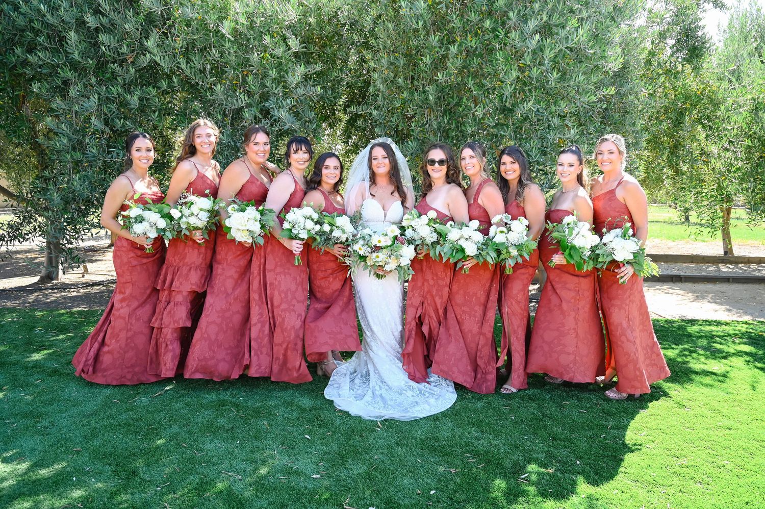 Bridal party in burgundy dresses and white gown pose on green lawn for group photos.