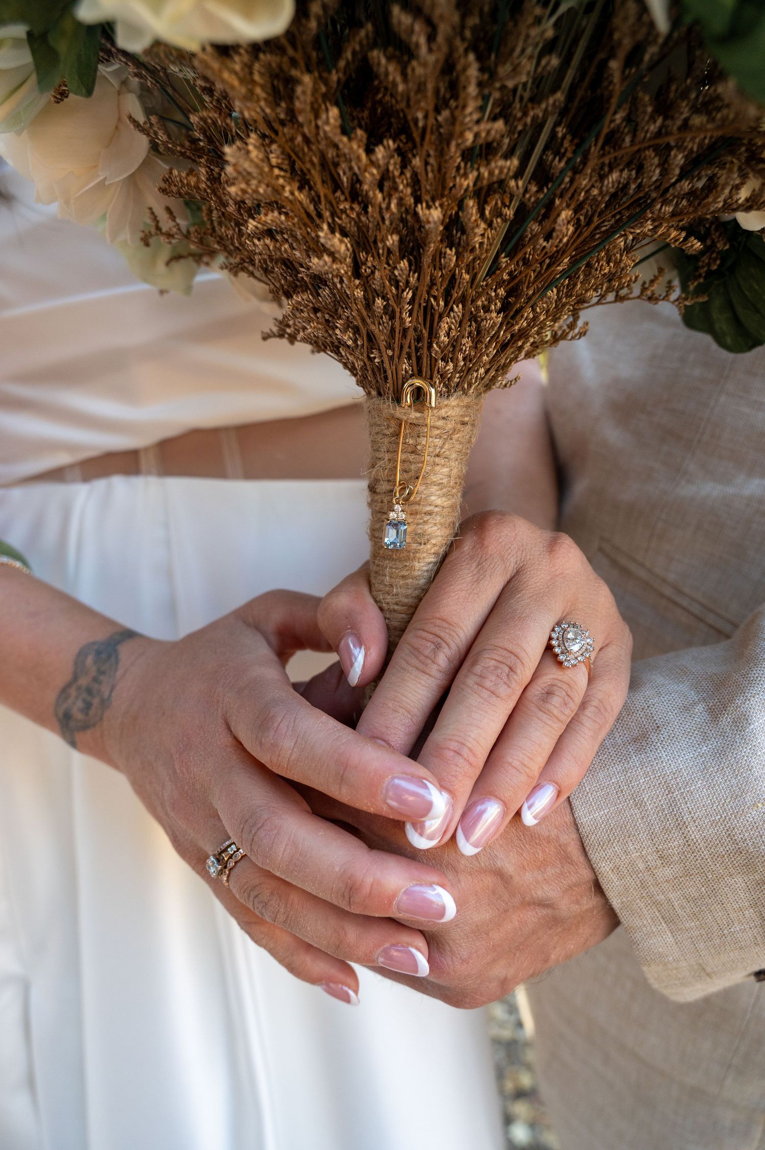 Carmel Valley Close-up view of hands with French manicure holding dried floral bouquet wrapped in twine against white fabric background.