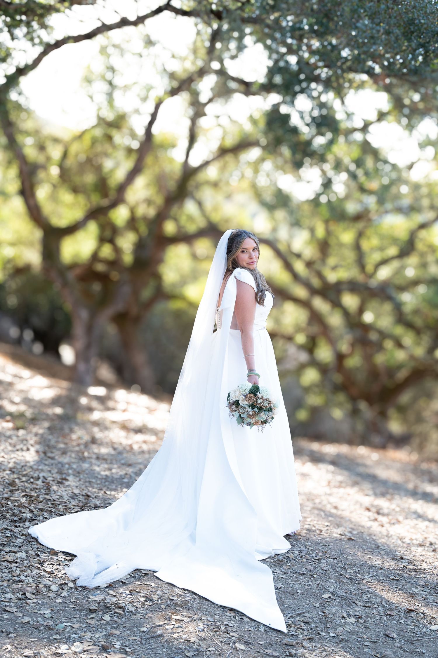 A bride in a flowing white wedding gown poses in a sun-dappled outdoor setting among oak trees.