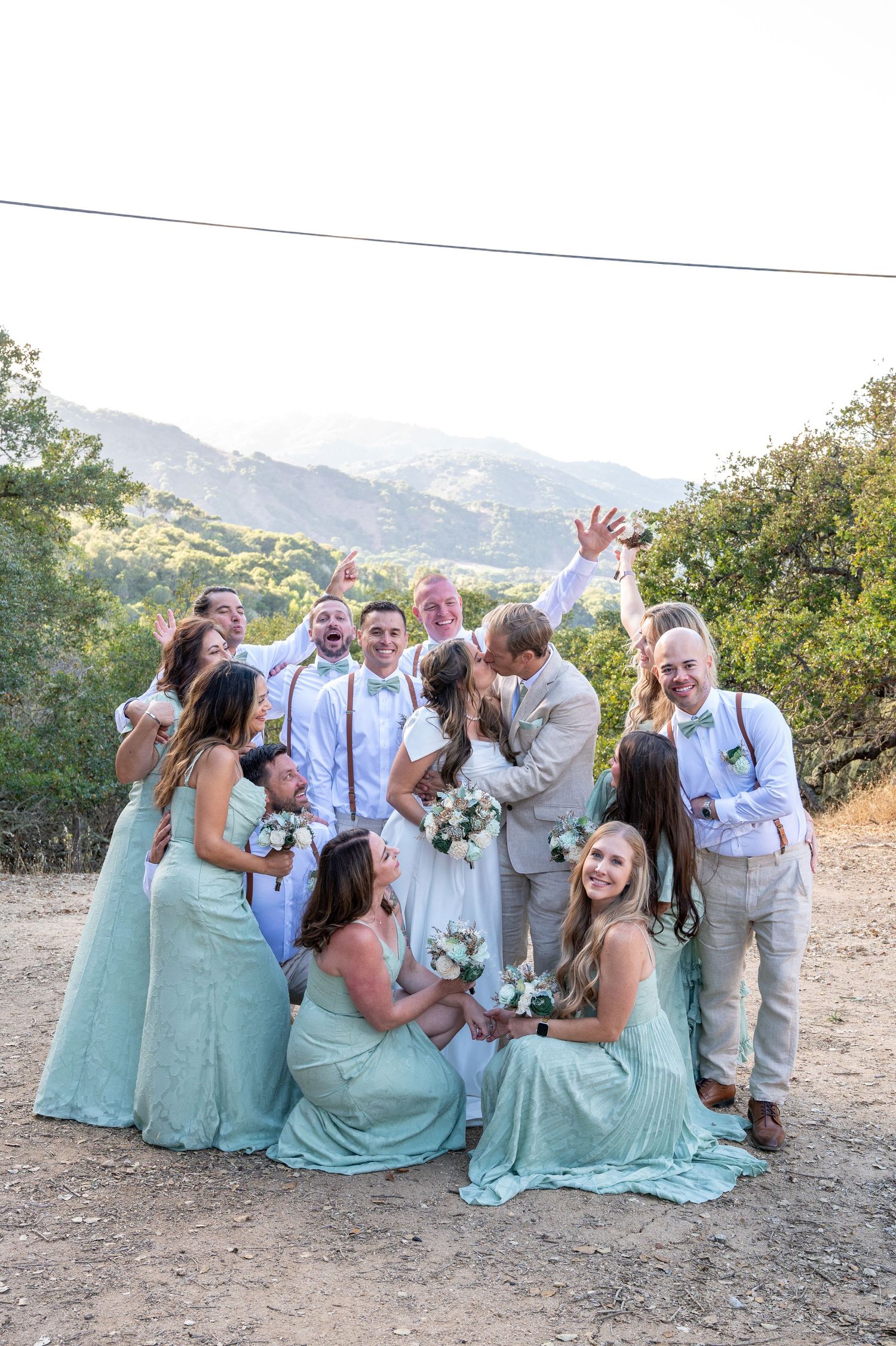 Wedding party in mint green dresses and gray suits pose playfully on a mountain trail with scenic views in background.