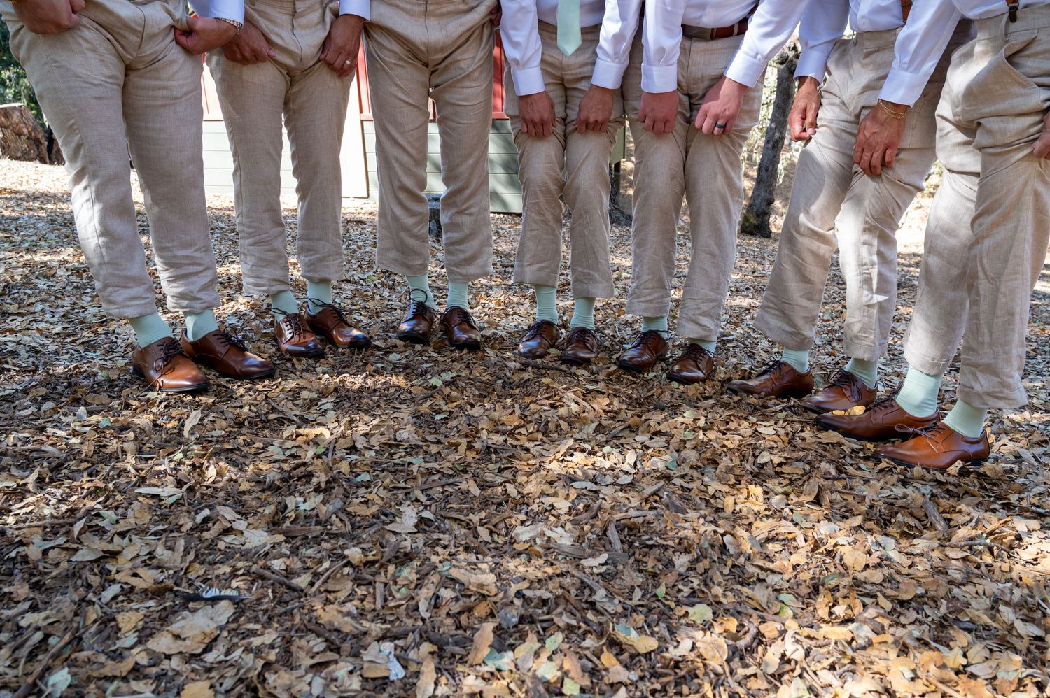 Multiple pairs of white shoes and feet standing in a line on dirt and leaves outdoors.