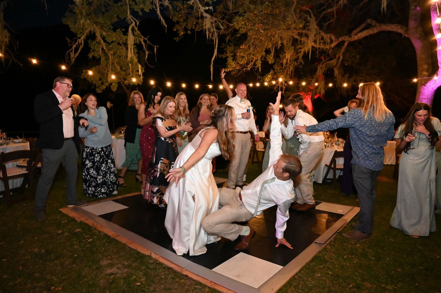 Wedding guests dance and celebrate on an outdoor wooden dance floor under string lights at a nighttime reception.