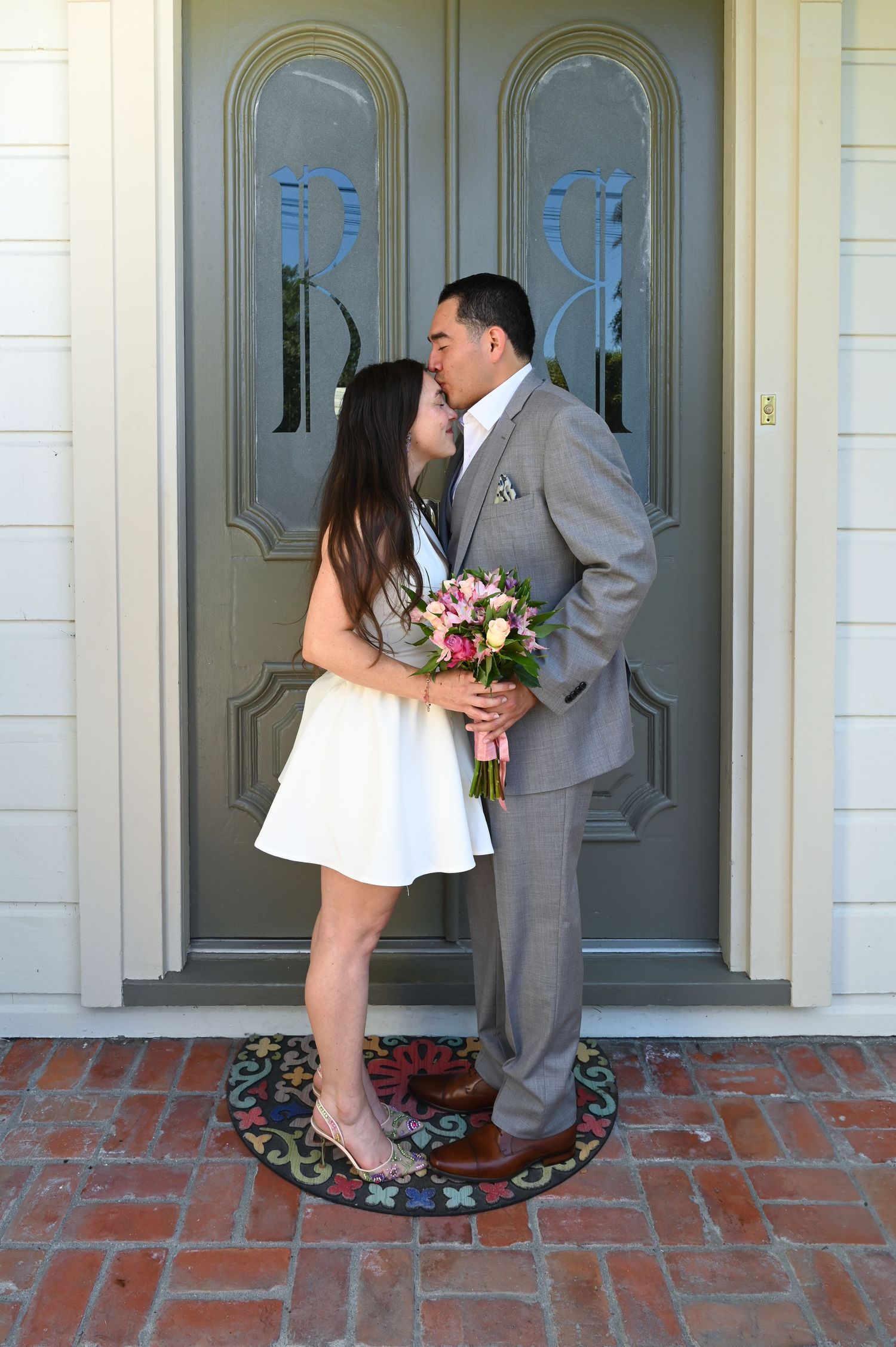 Wedding couple stands on brick porch in front of classic white doorway.