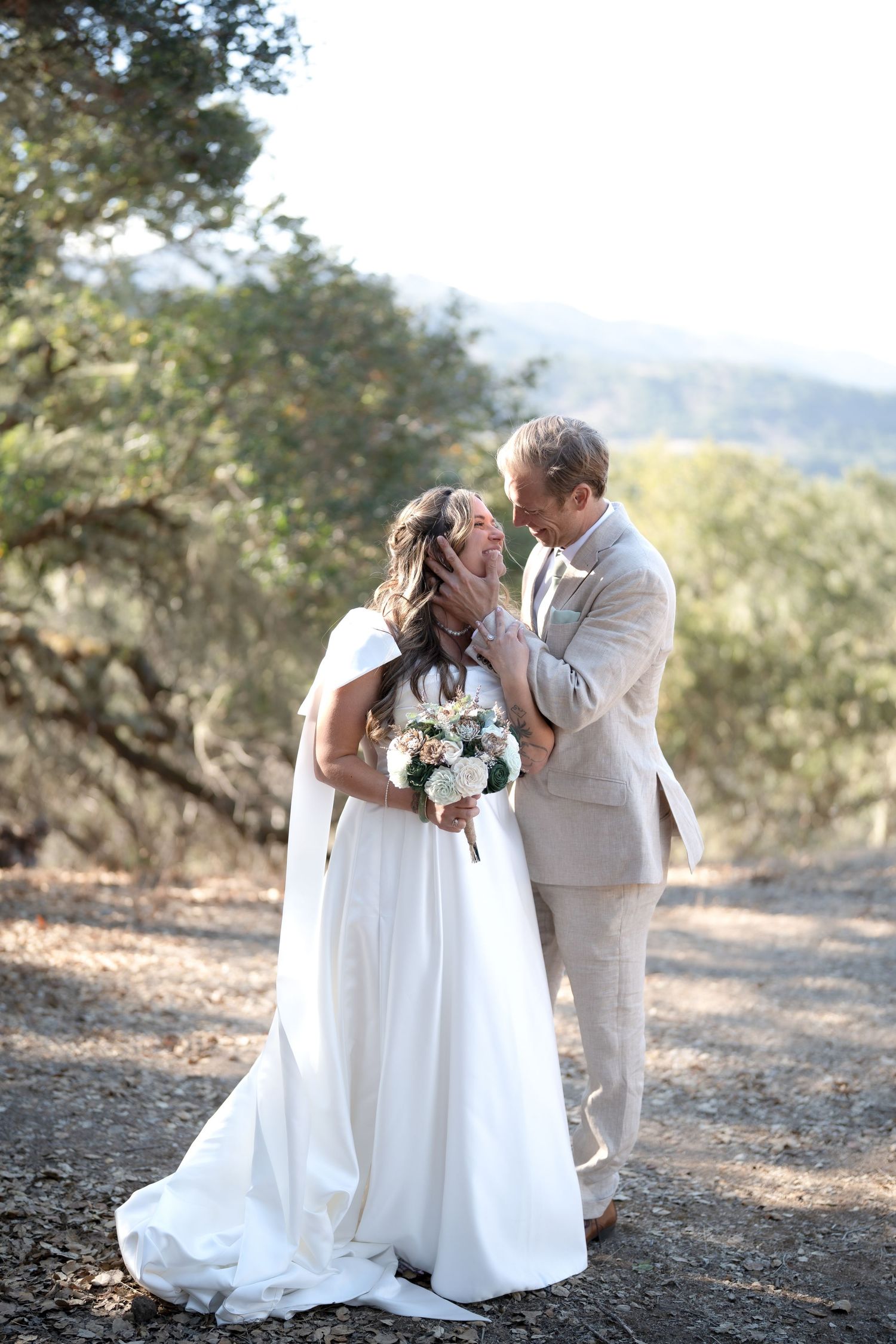 Carmel Valley Wedding, California Two people in white wedding attire share an intimate moment outdoors against a natural mountain backdrop.