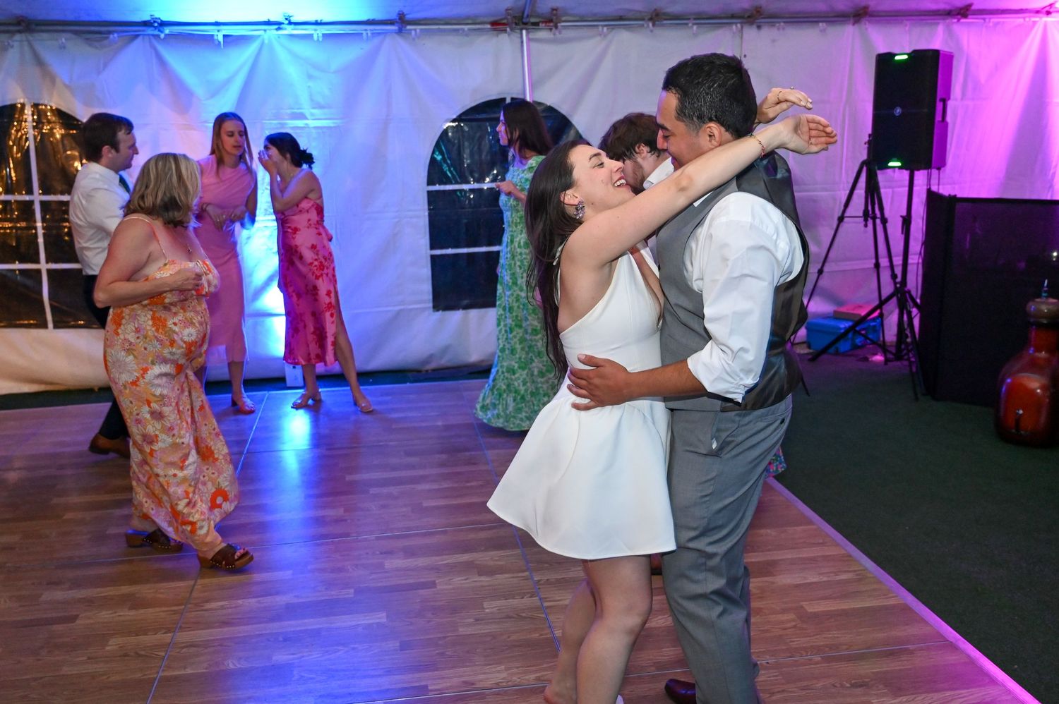 Wedding guests dancing under purple mood lighting in a decorated reception tent.