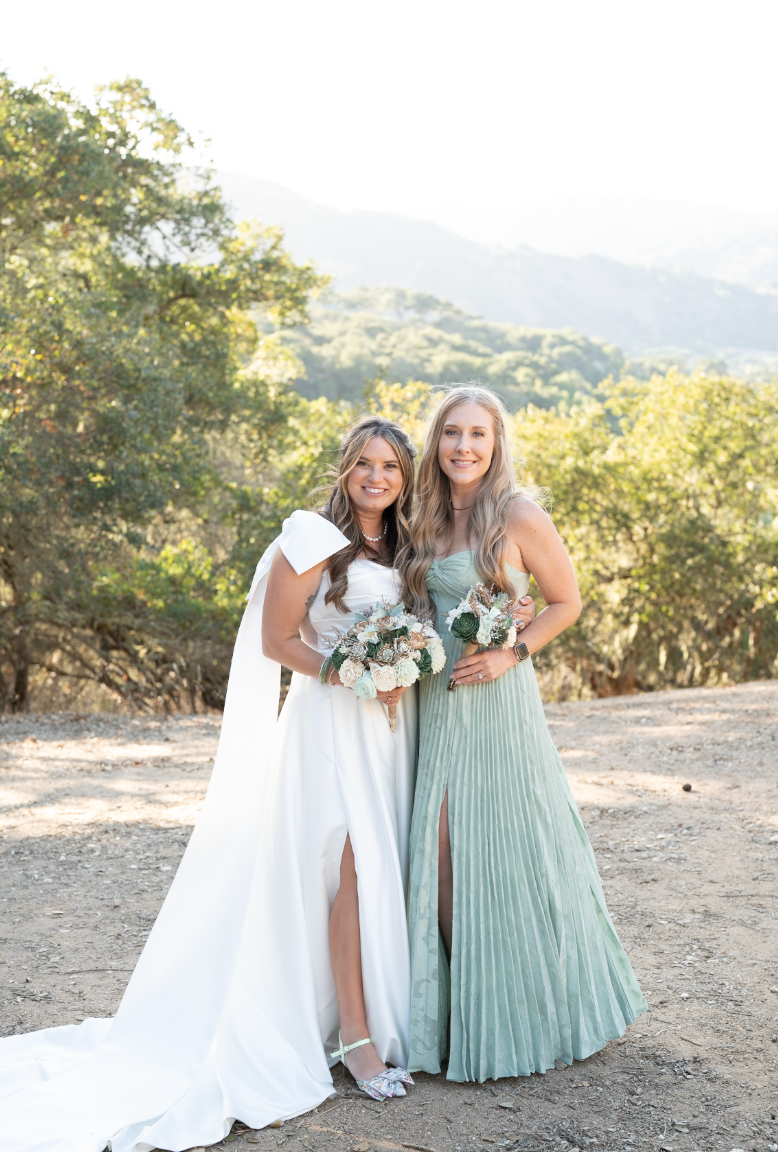 Three people in elegant dresses laugh and embrace outdoors on a sunny day with mountains in the background.