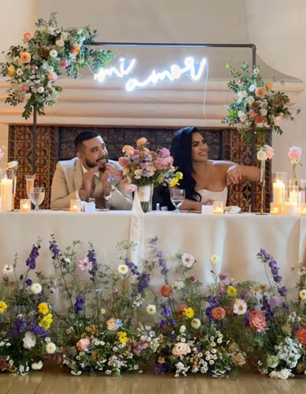 Wedding reception venue decorated with lush floral arrangements and candles along a white wall display with neon signage.