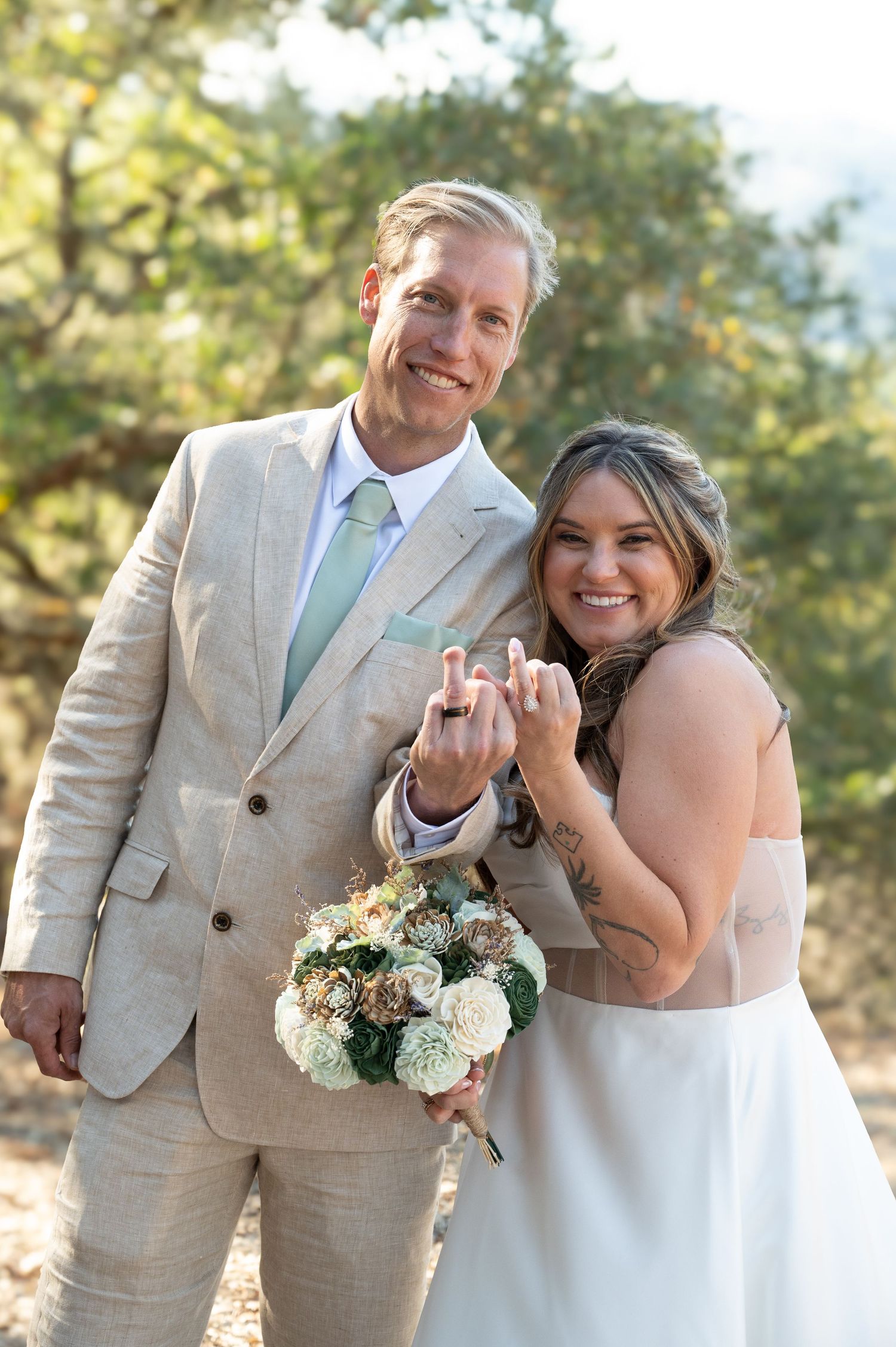 Wedding couple in tan suit and white dress showing off rings with joyful expressions.