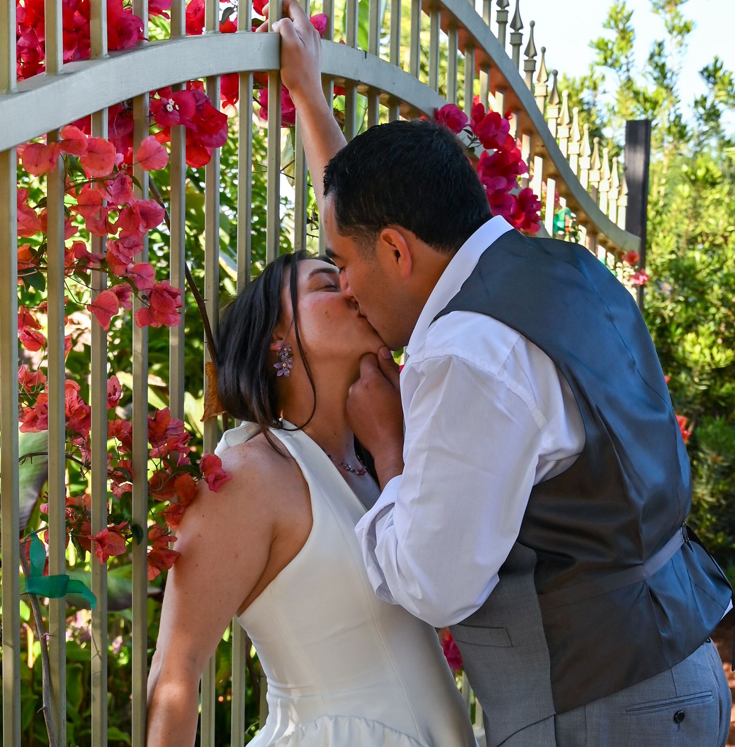 A couple sharing a romantic kiss near a fence decorated with red climbing flowers.