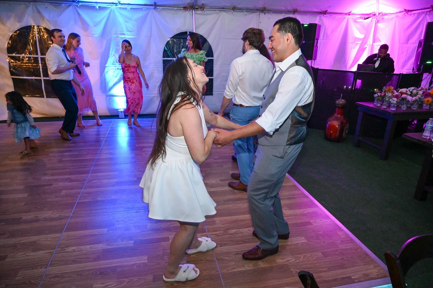 Wedding party dancing on wooden floor under purple and pink lighting in tent venue.