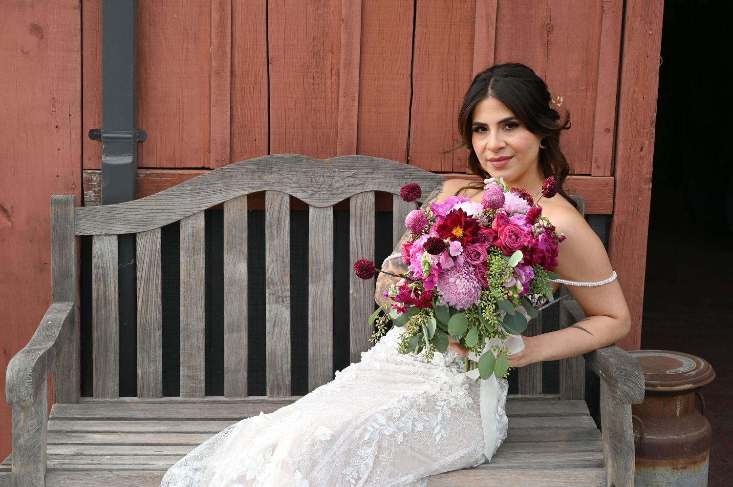 A bride in a lace dress poses with a vibrant pink and burgundy floral bouquet against a rustic red barn backdrop.