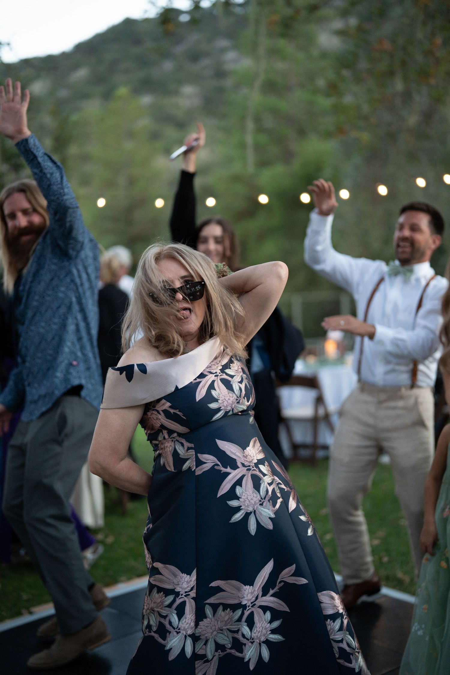 People dancing and celebrating at an outdoor party with string lights and trees in the background.