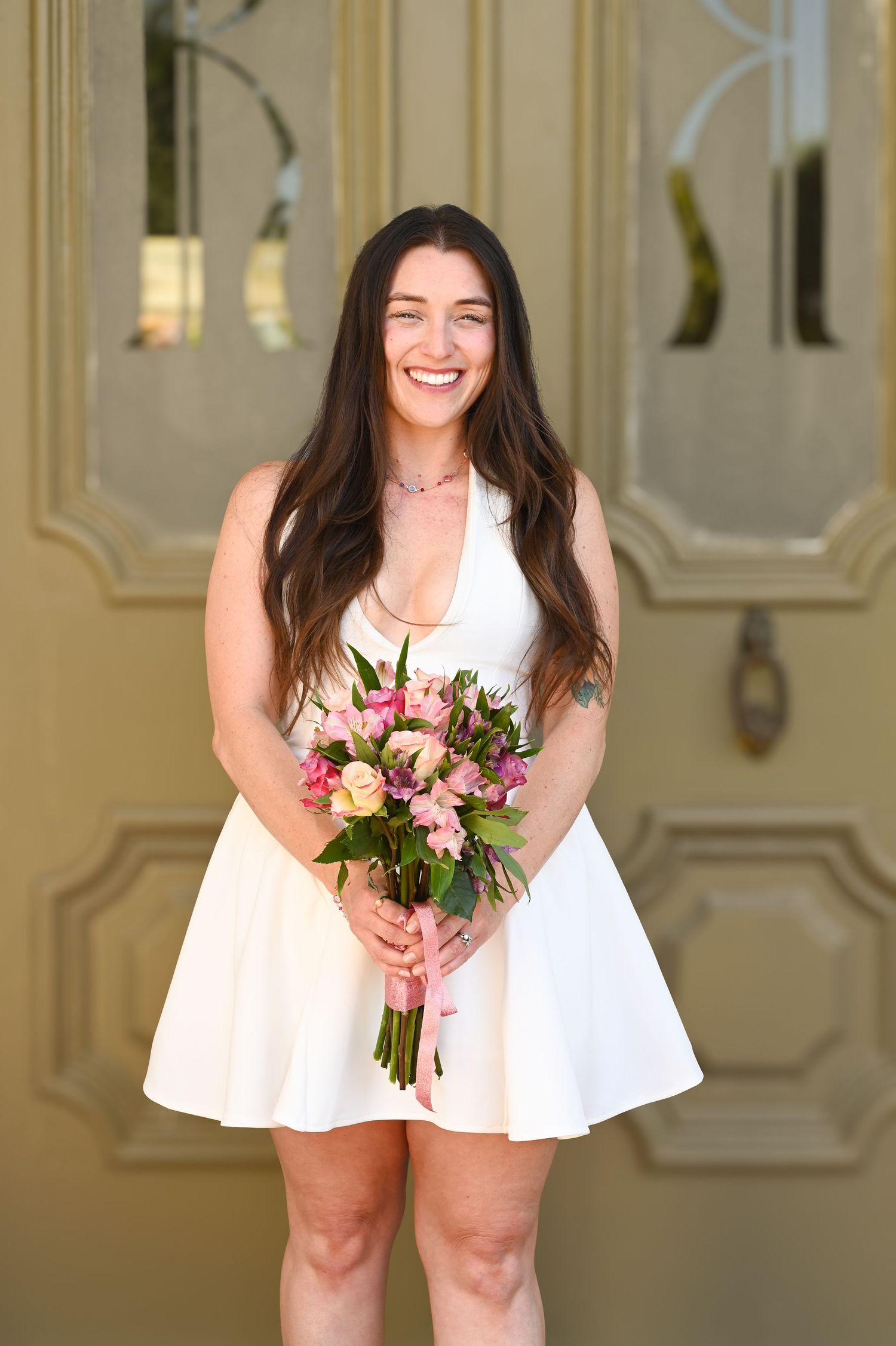 Bride in white dress holding pink and purple floral bouquet in elegant indoor setting.