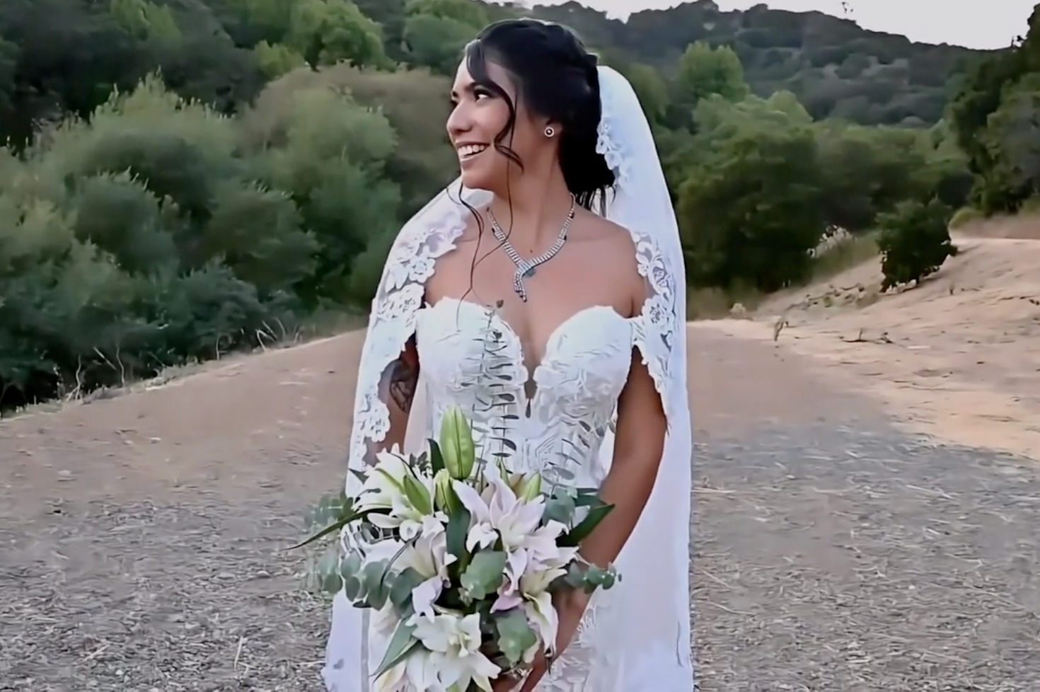 A bride in a white lace wedding dress holds a delicate bouquet of white and pink flowers against a desert landscape.