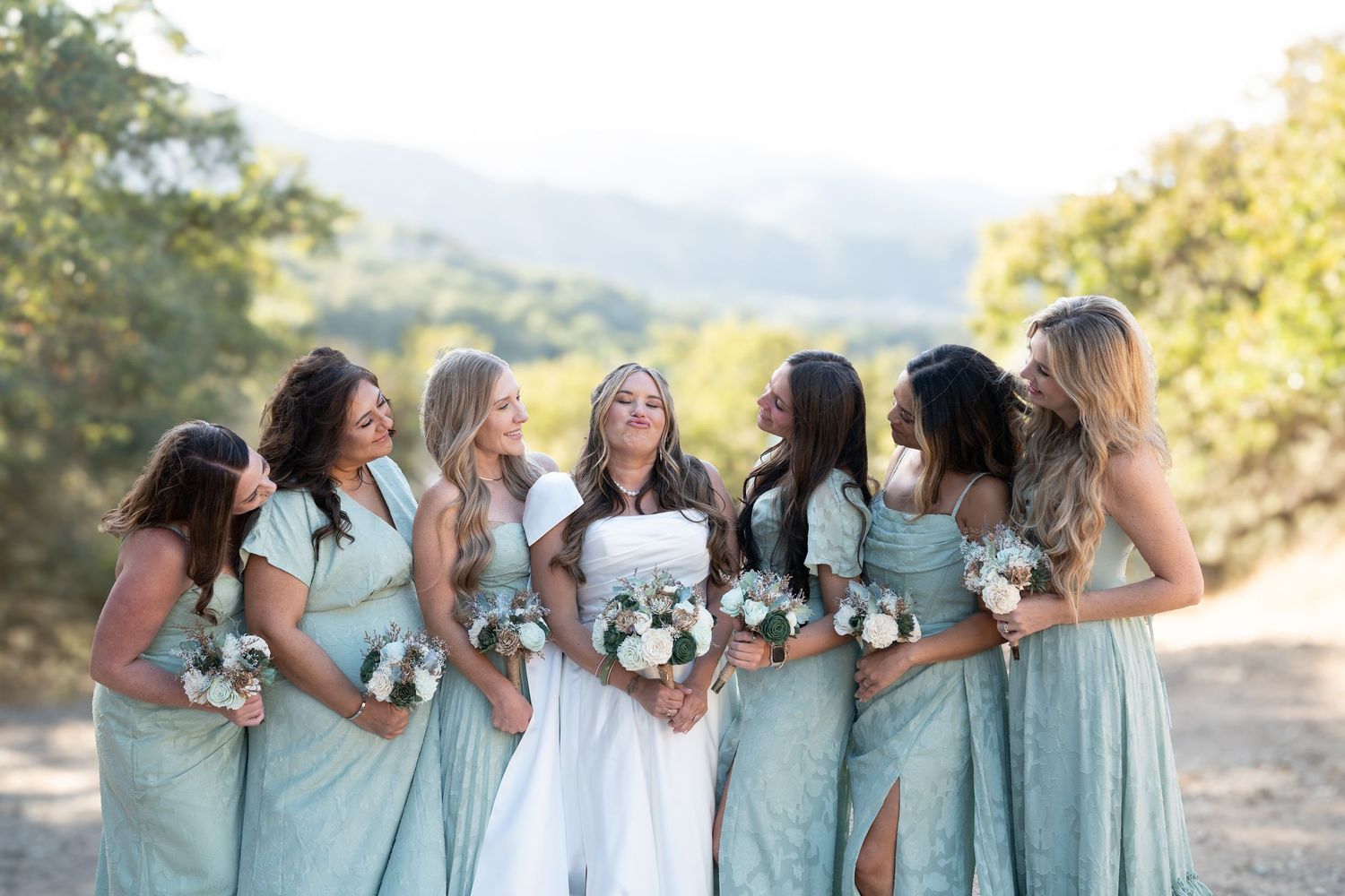 Bridesmaids in matching mint green dresses stand together holding bouquets during an outdoor wedding photoshoot.