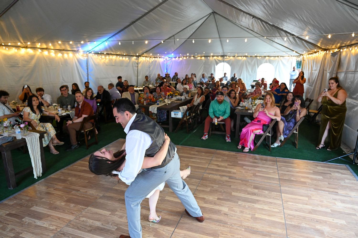 Large wedding reception tent with string lights and guests dancing on wooden flooring.