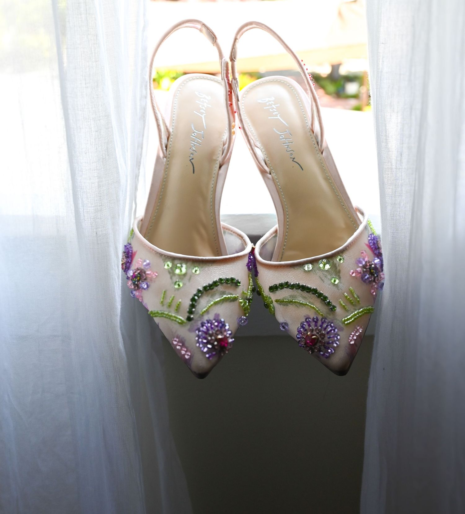 Delicate floral-decorated bridal shoes displayed against a white curtain background.