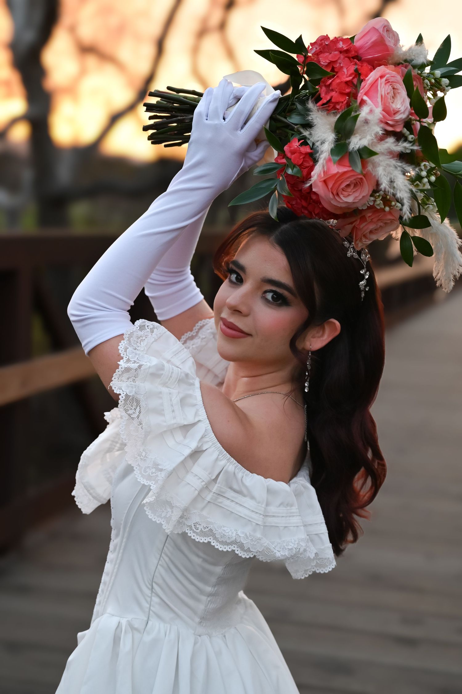 Bride in off-shoulder dress poses dramatically with bouquet at sunset on wooden deck.