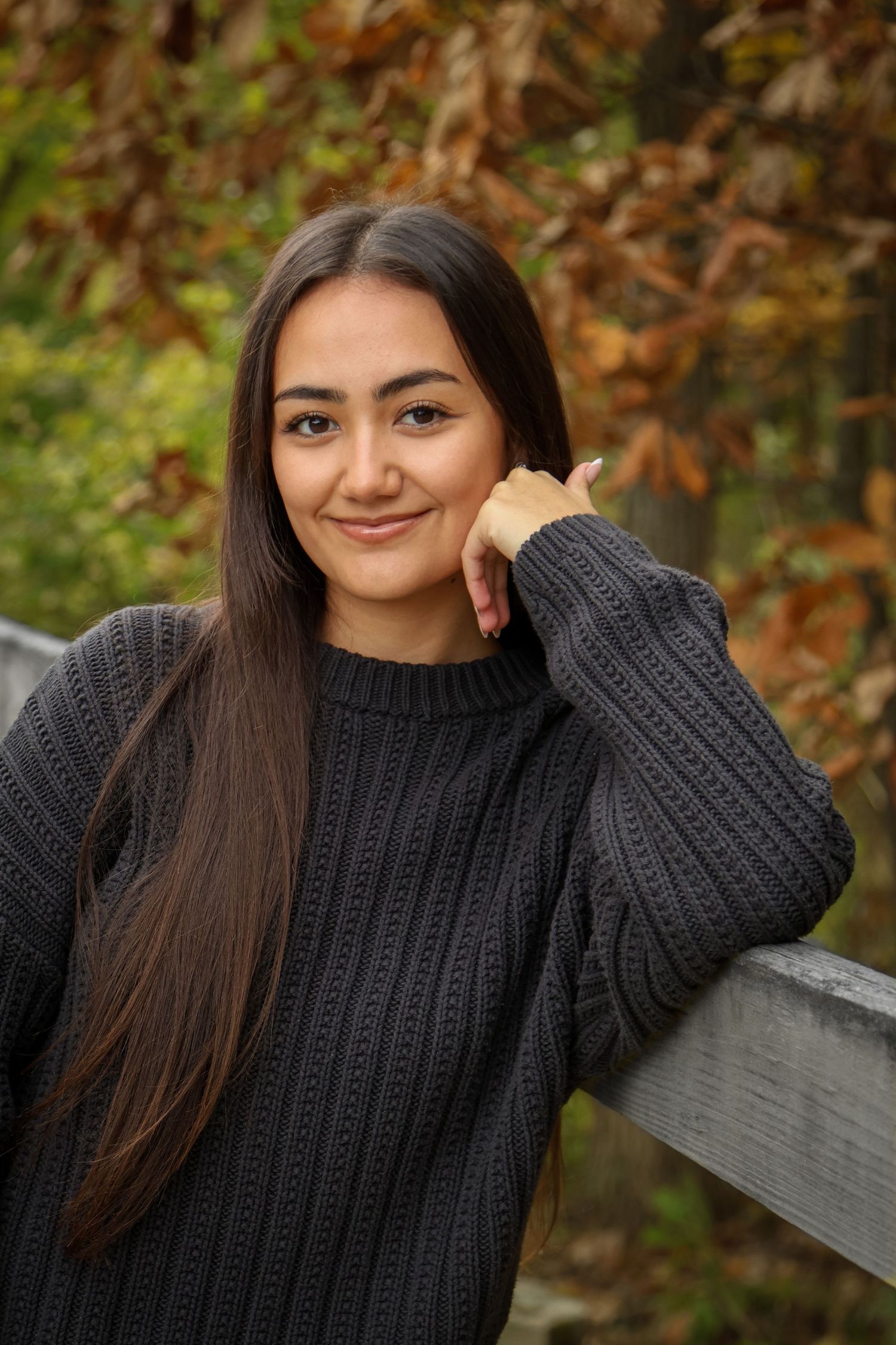 Senior girl gently smiles at camera while her hand gently props up her face and she is surrounded by fall foliage