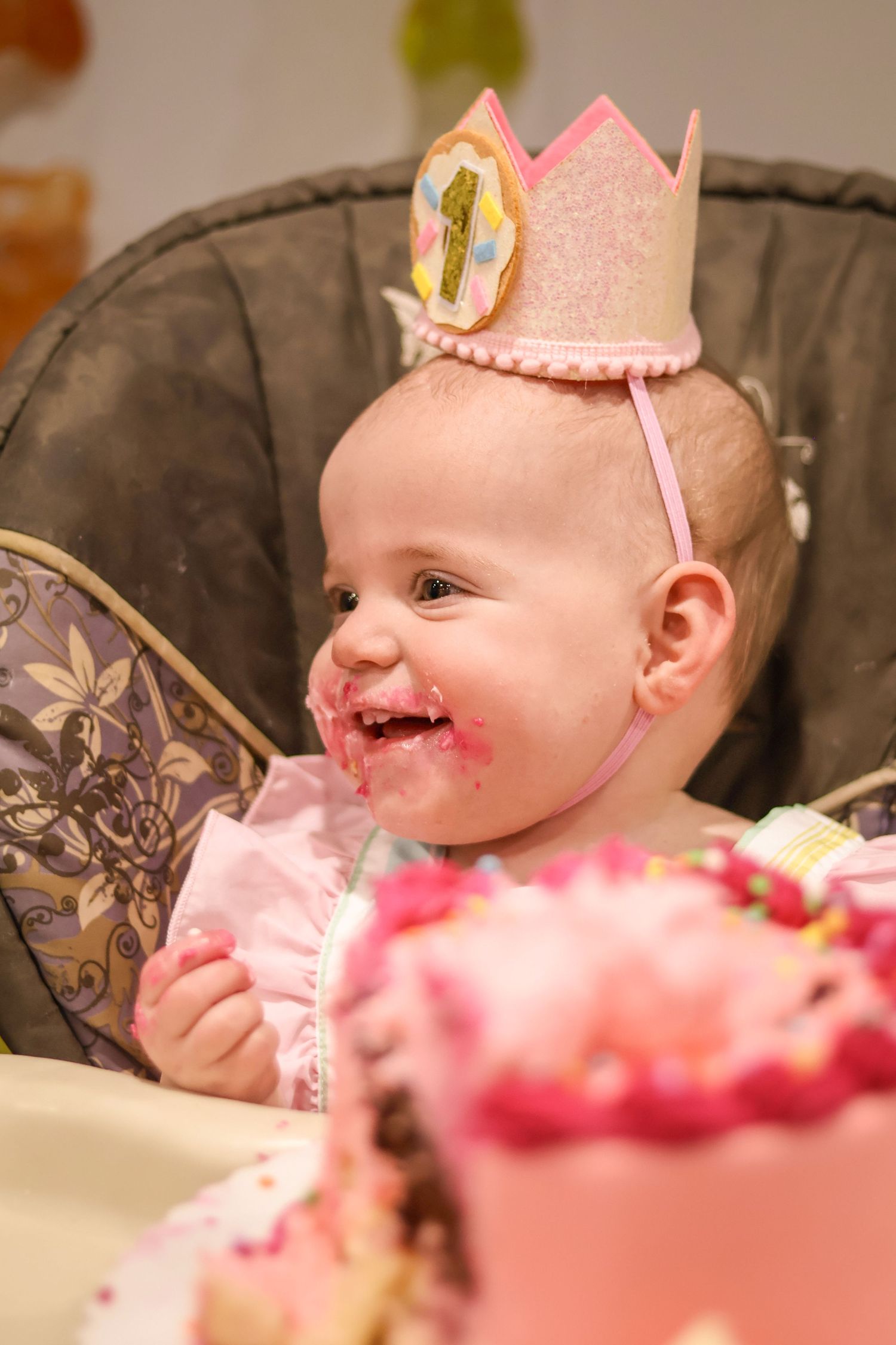 One year old girl smiles as she wears a birthday crown. Pink cake in the foreground