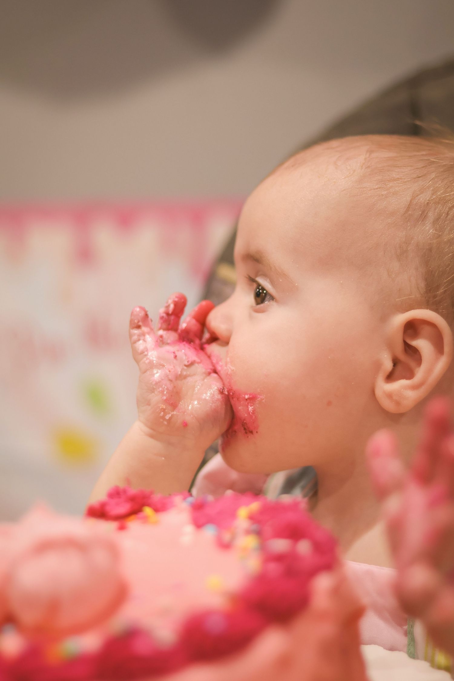 One year old girl eats cake frosting off her fingers. Pink cake in the foreground