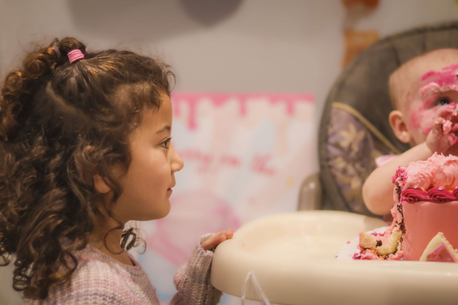 Cake smash with cousin looking on as babies face is covered in pink frosting