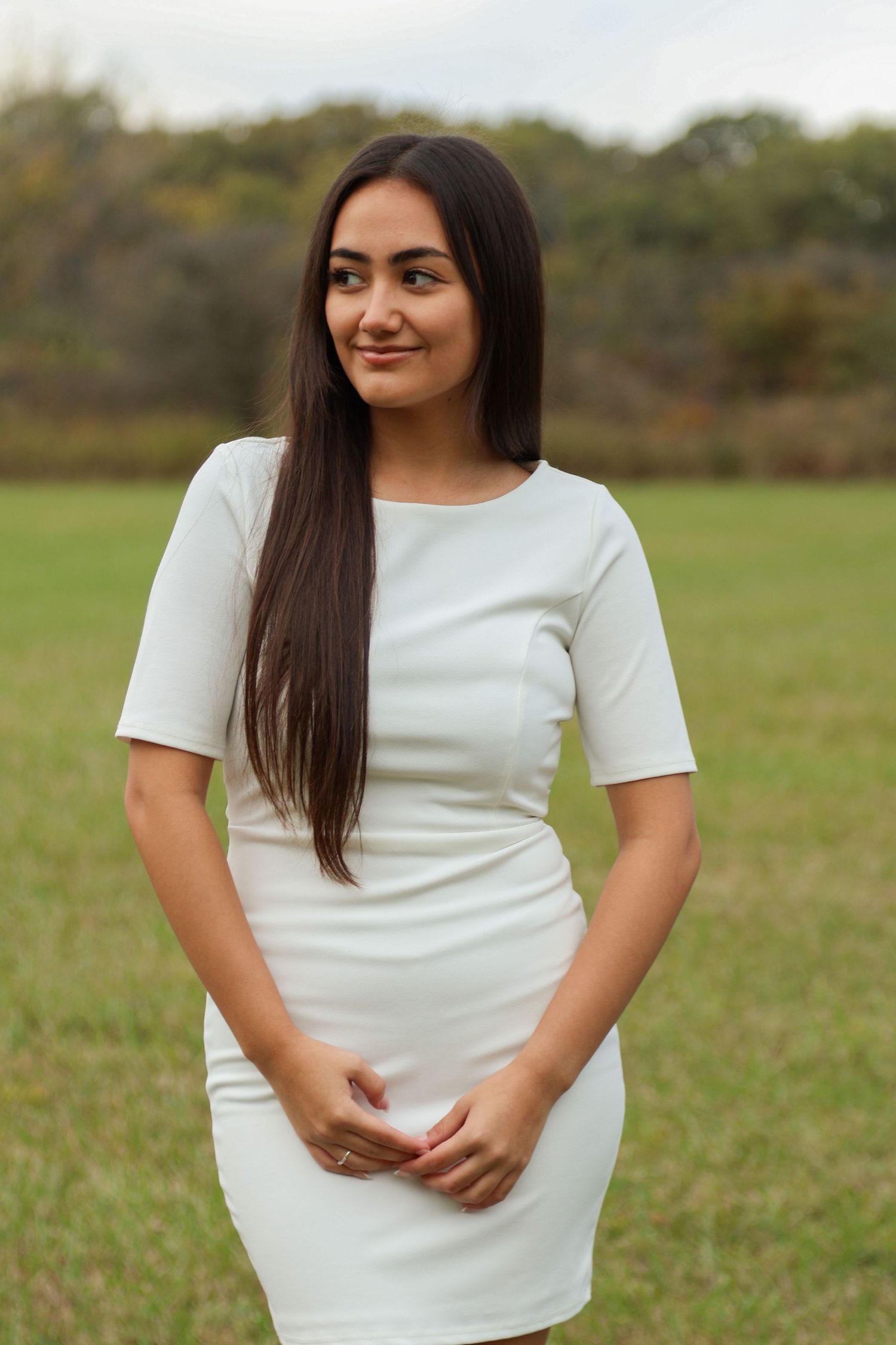 Senior girl wears white dress in a field with fall foliage and has gentle smile on her face