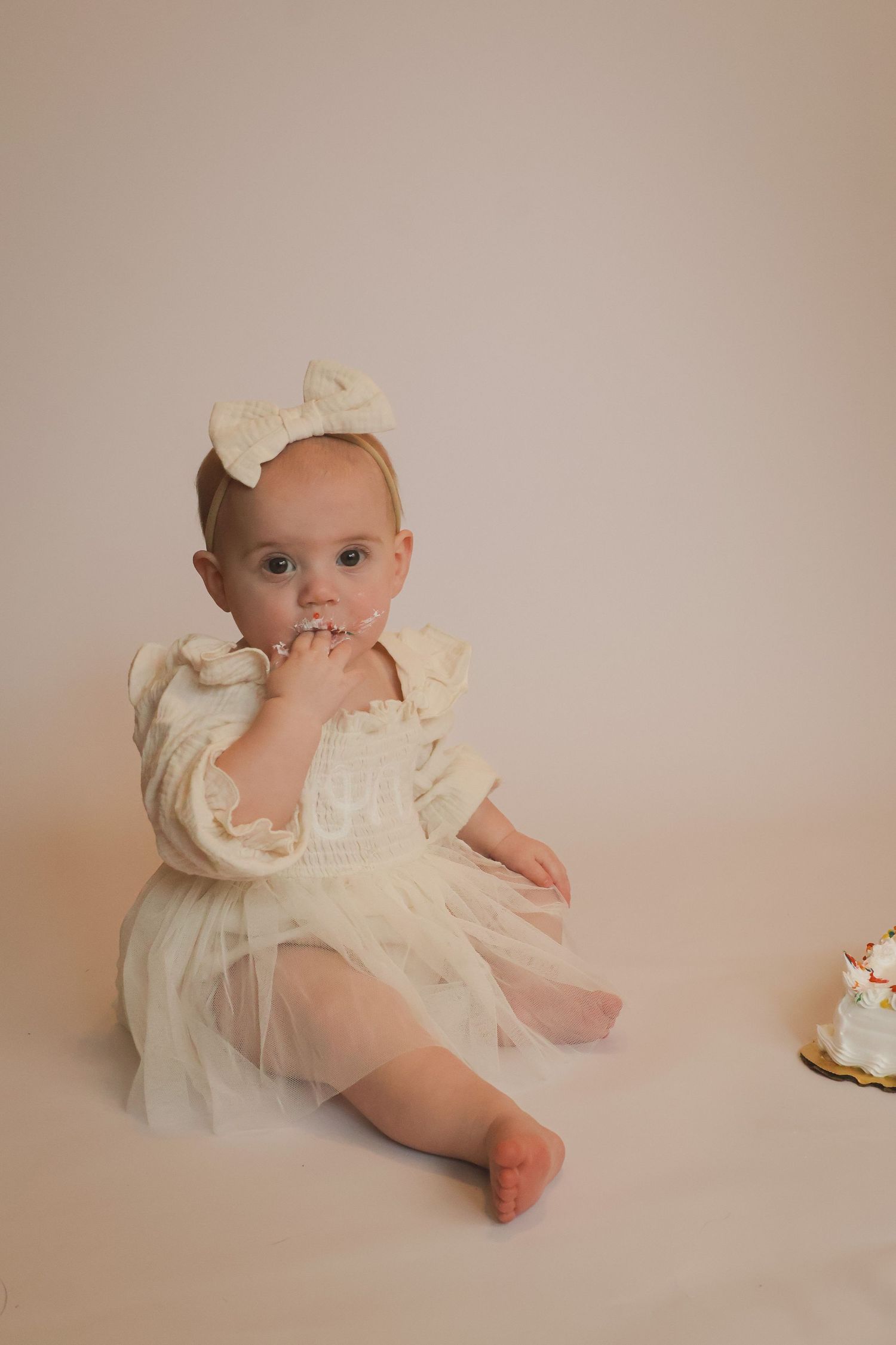 One year old baby girl in a cream dress and bow headband enjoying cake during a studio cake smash session by Kristin Photo Co in Beecher IL