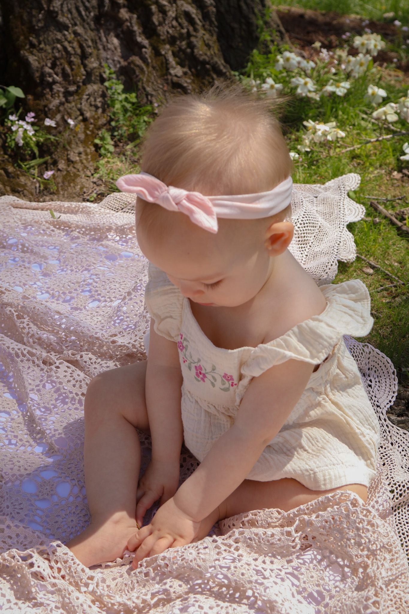 One year old girl sits on a lace blanket with a cream and baby pink bow and outfit photo