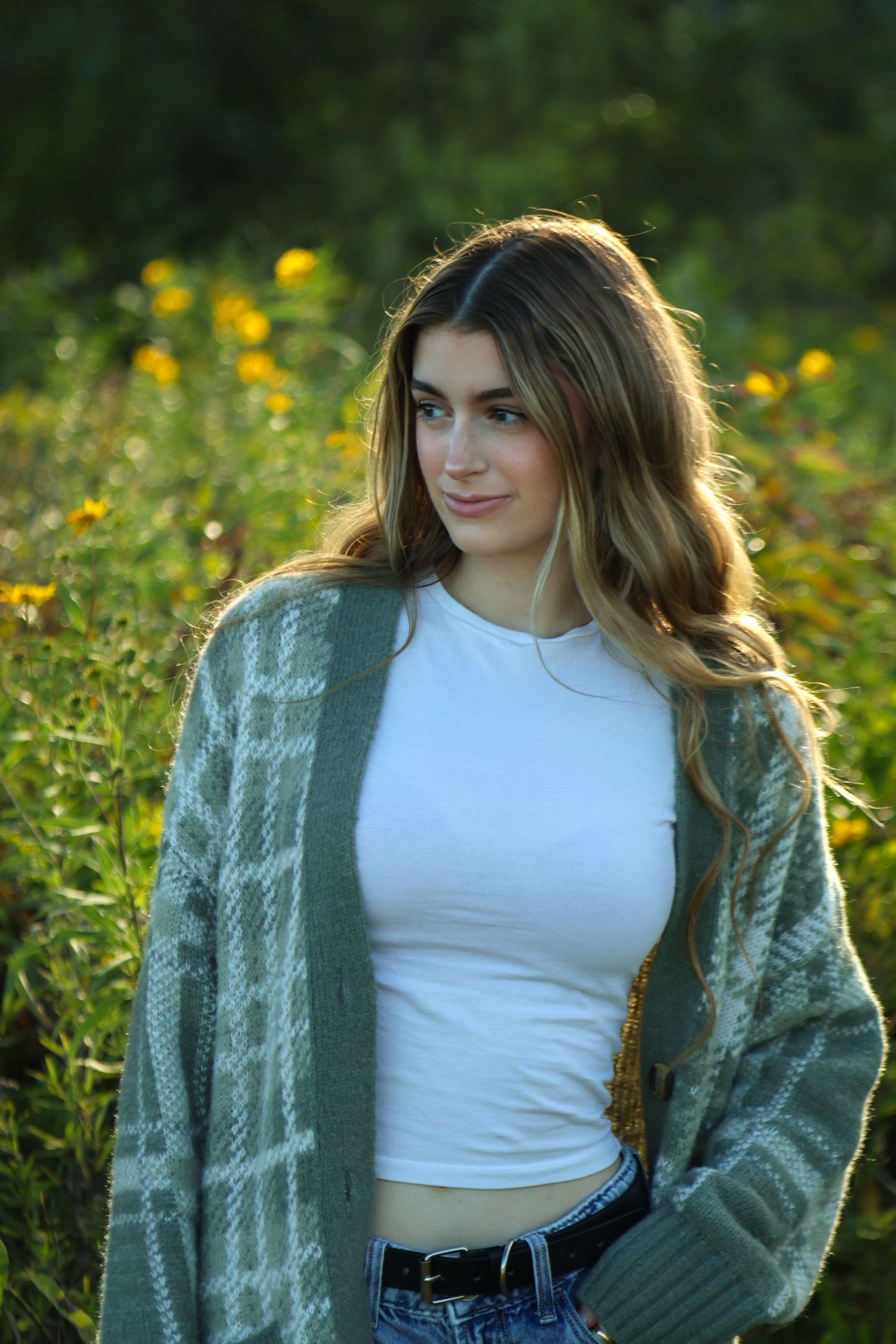 Senior girl in cozy green sweater with beautiful yellow flowers behind her white she looks to the side and has a coy smile