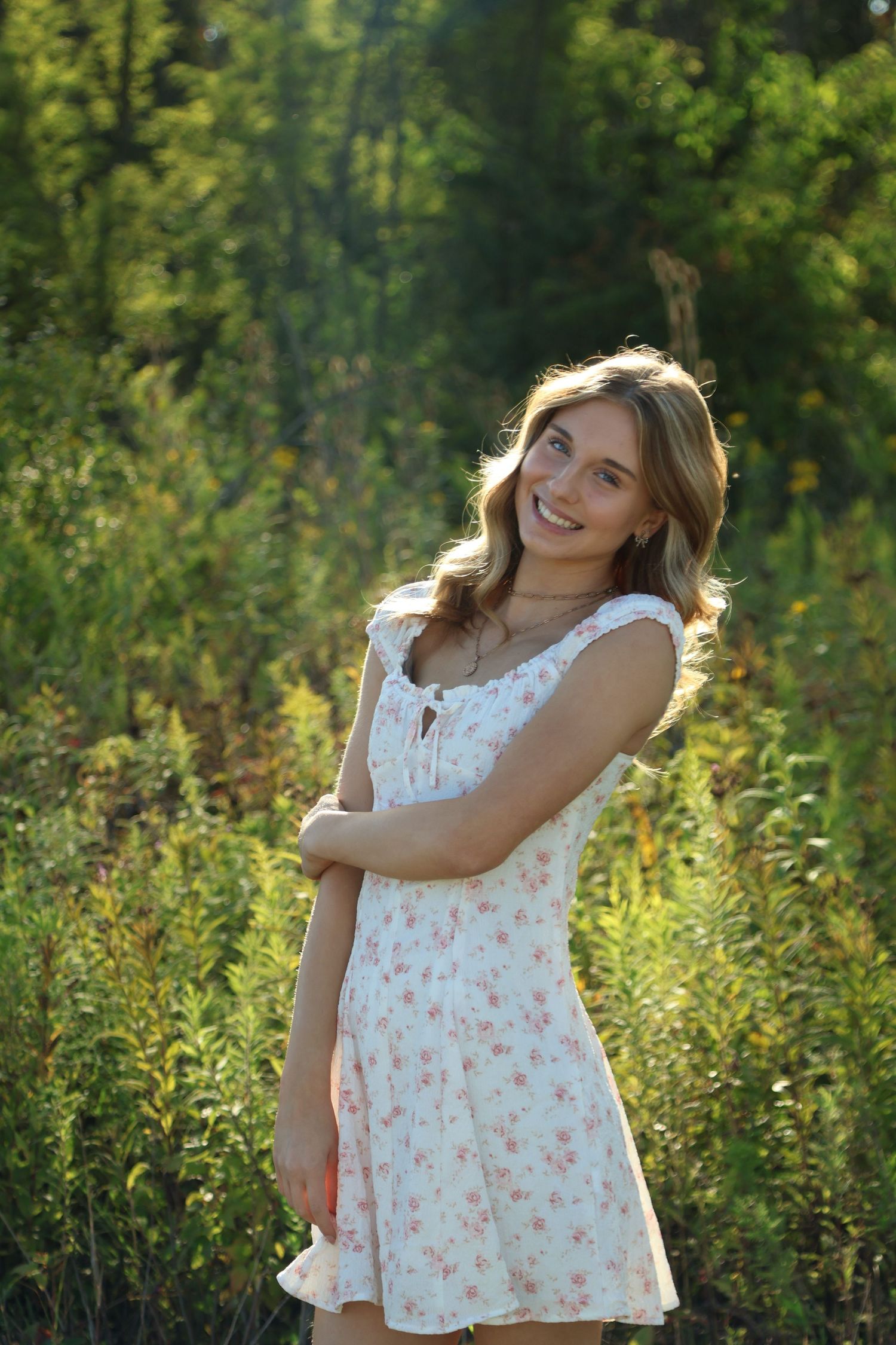 Blonde senior girl is wearing a white floral dress and surrounded by bright green foliage while she smiles and holds her arm in sweet pose