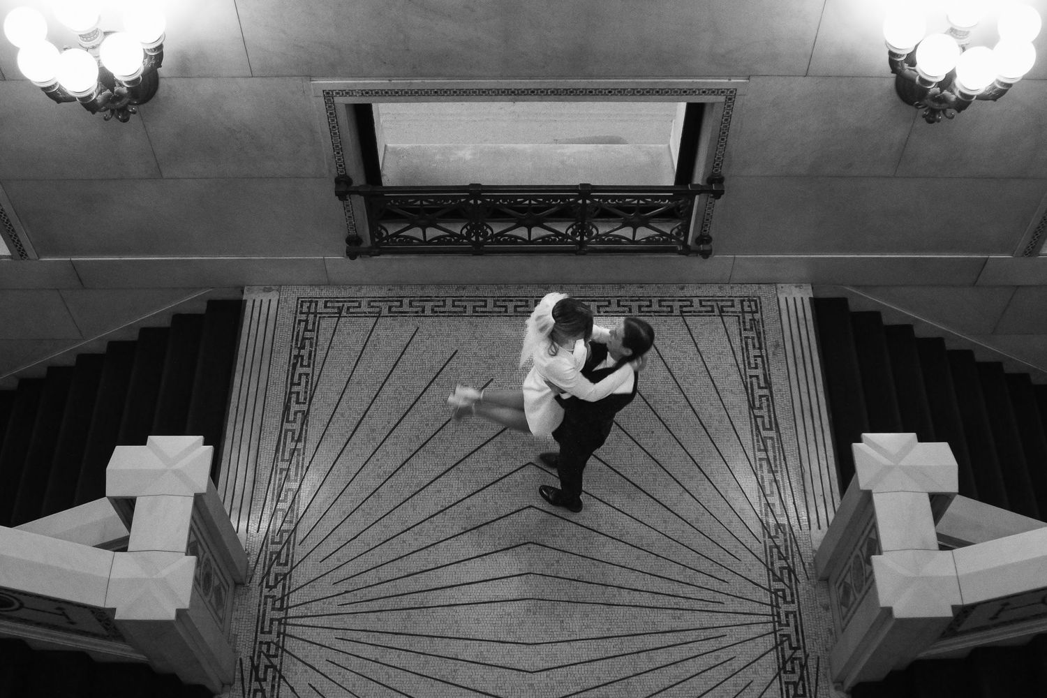Black and white overhead photograph of a bride and groom spinning together on a tiled floor. Her dress flaring softly as the hold each other
