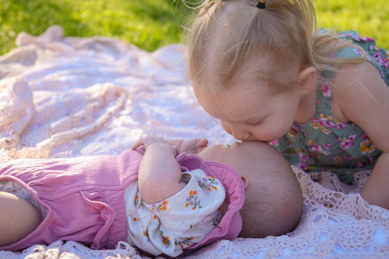 Toddler sister kisses baby sisters head on a blanket in the spring with sunlight basking on them taken in grant Park IL