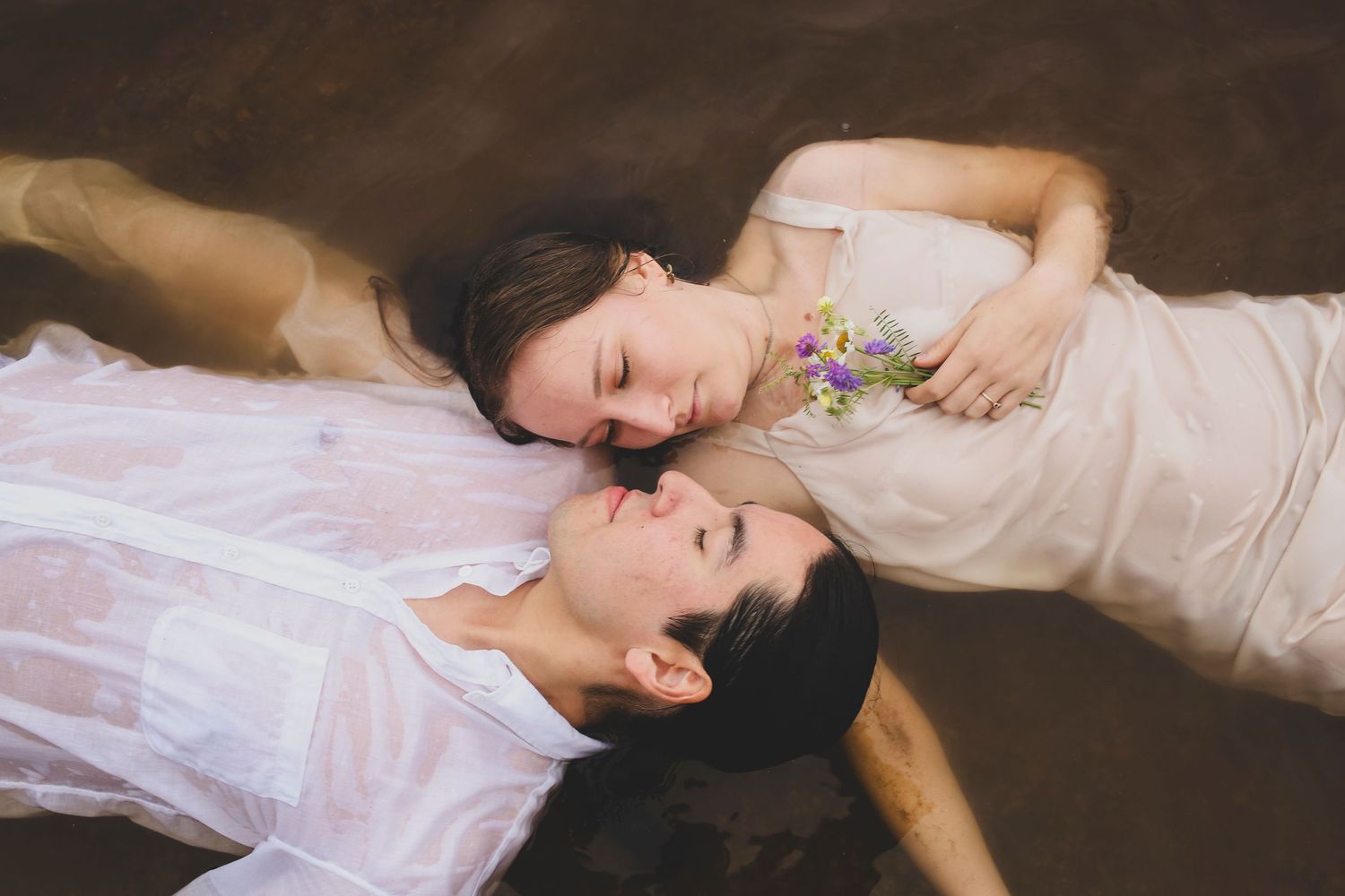 Two women in white dresses lying in shallow water, one holding wildflowers, in an artistic portrait.