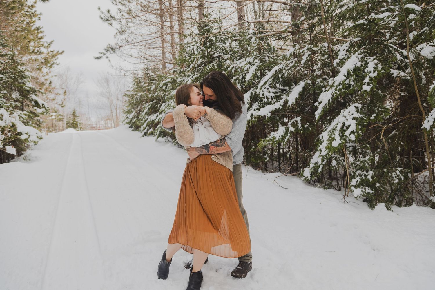Two women embracing joyfully on a snowy forest road in winter, surrounded by snow-covered evergreen trees.