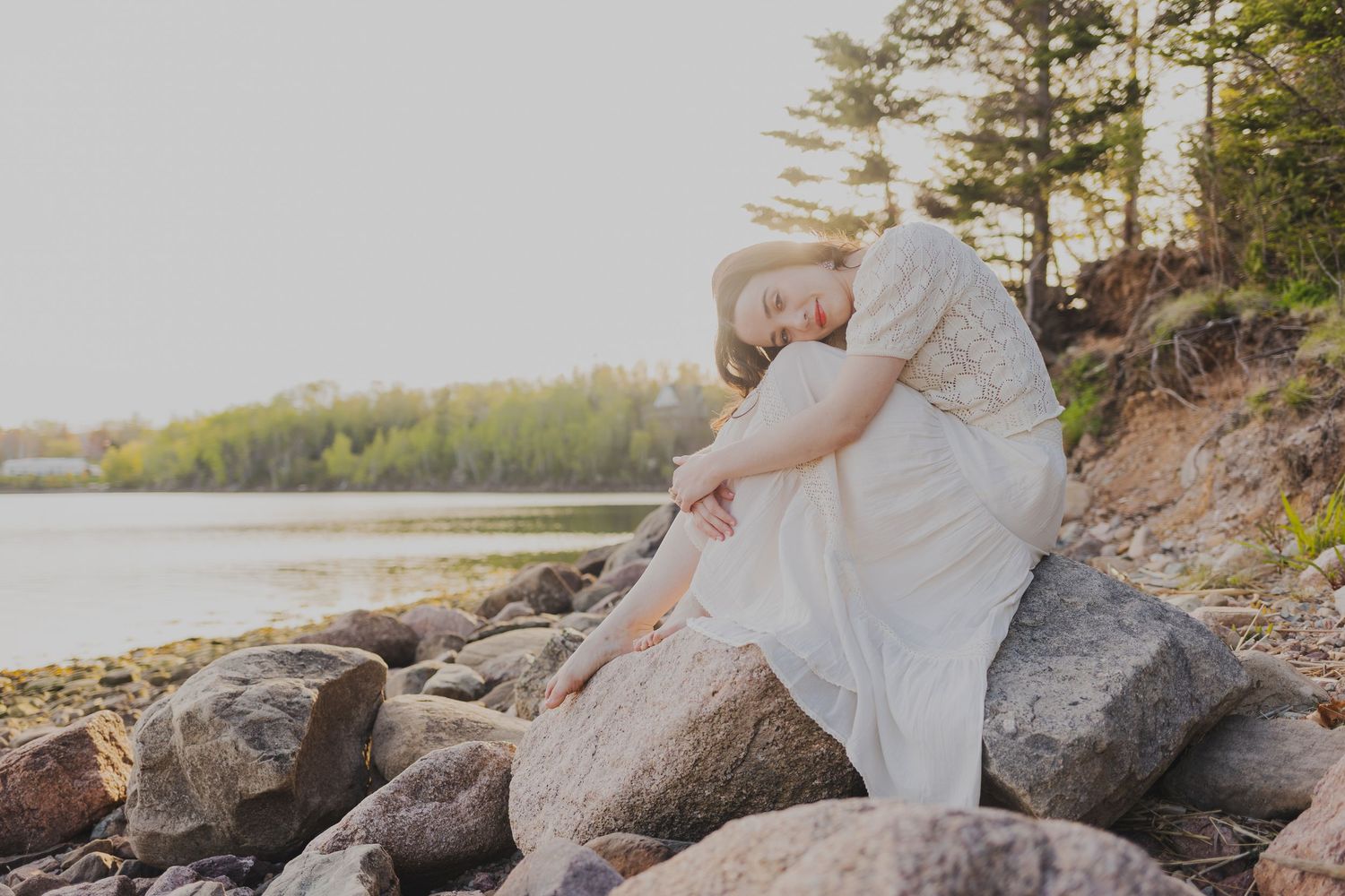 A young woman in a white dress sits peacefully on rocky shoreline surrounded by trees and calm water at golden hour.