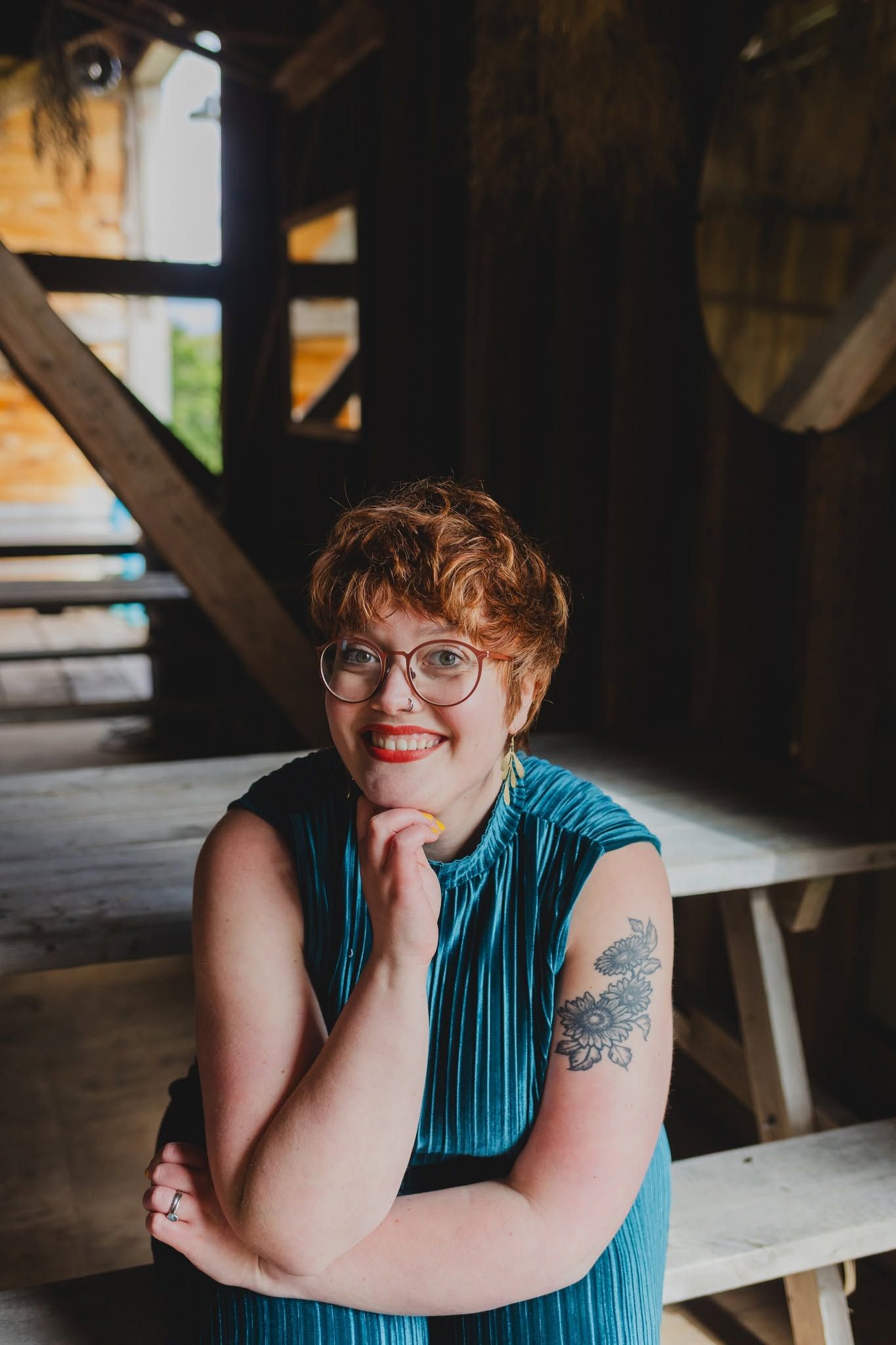 Smiling redhead woman with glasses and floral tattoo in teal dress seated in rustic wooden barn setting.