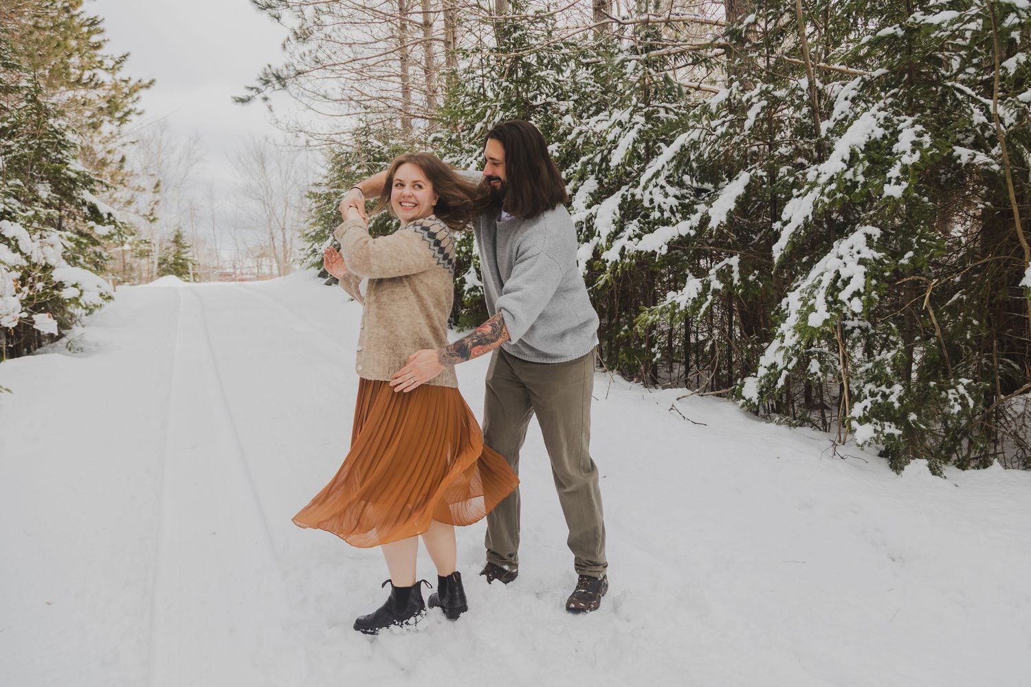 Couple dancing joyfully on a snowy forest road in winter, woman in rust skirt, man in gray sweater.
