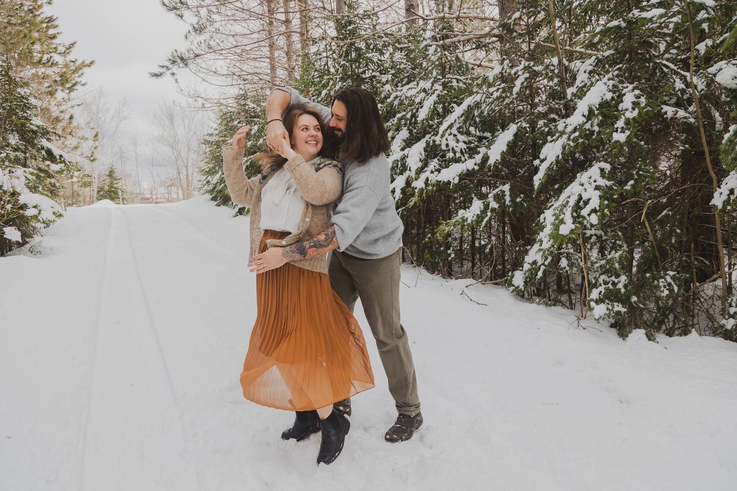 Couple dancing joyfully on a snowy forest road in winter, woman in orange skirt and man in gray outfit.