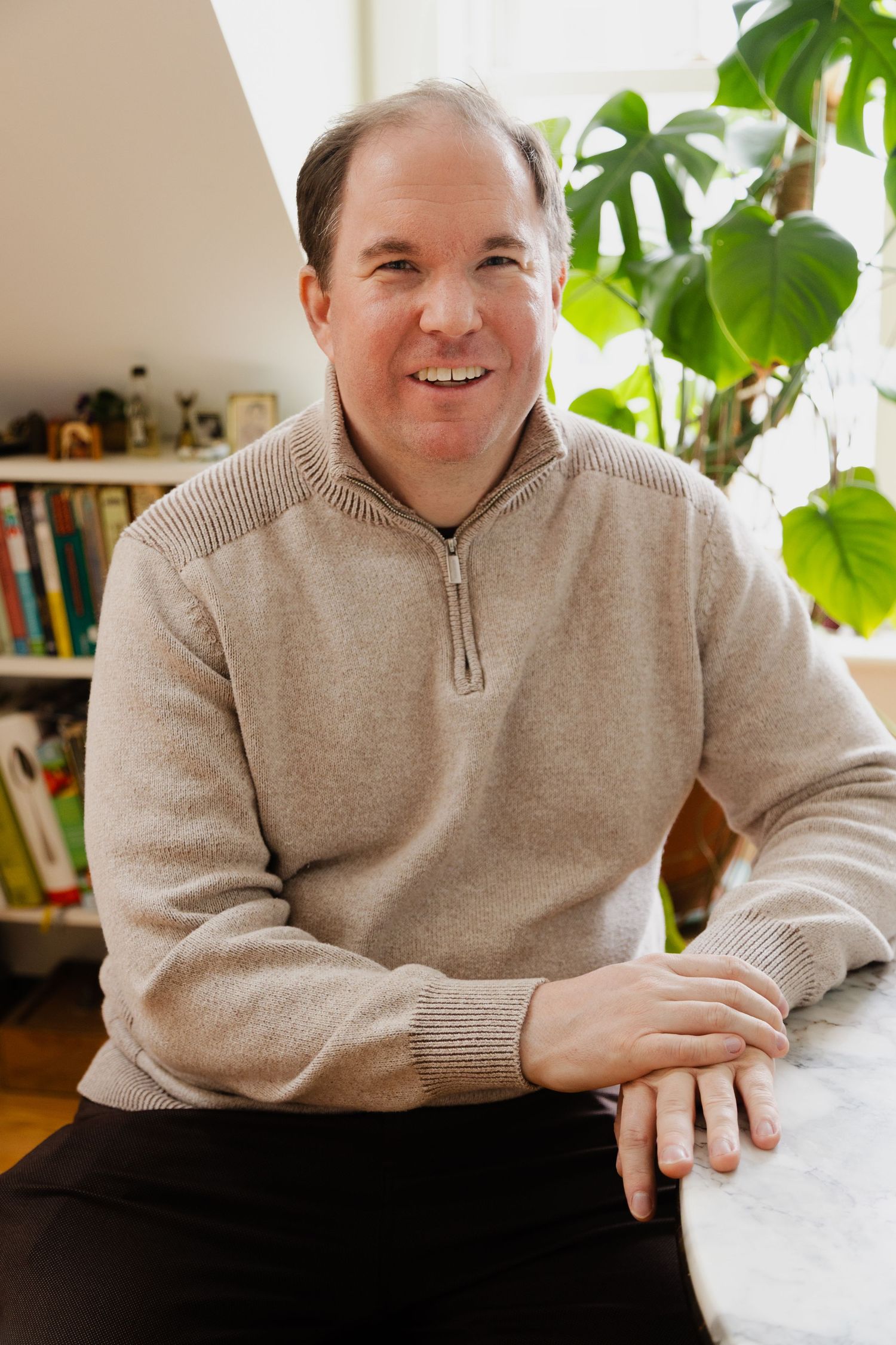 Portrait of someone in a beige sweater sitting on a couch with houseplants in the background.