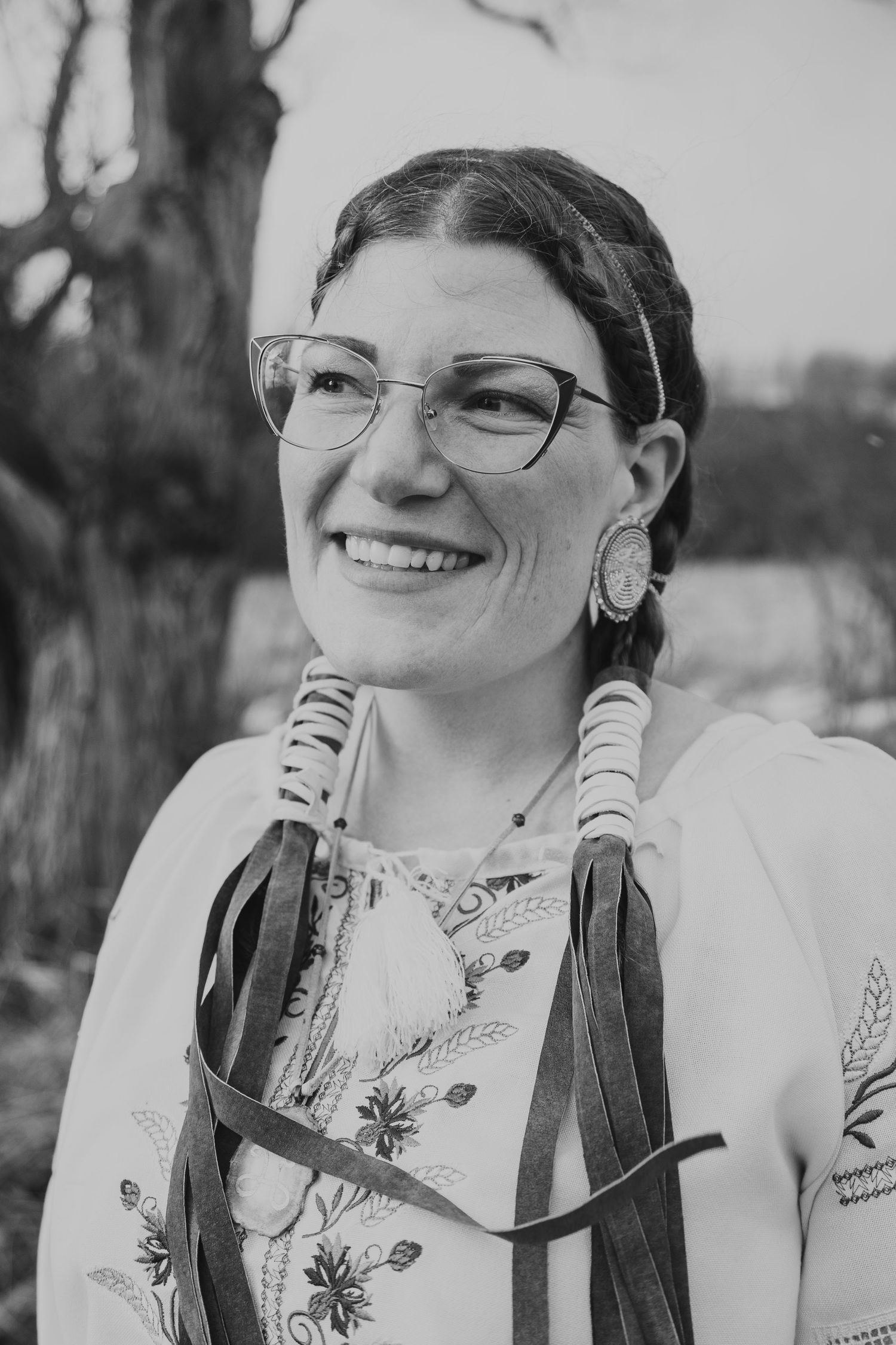 Smiling woman wearing glasses, beaded earrings, headband, and fringe necklace in a black and white outdoor portrait.