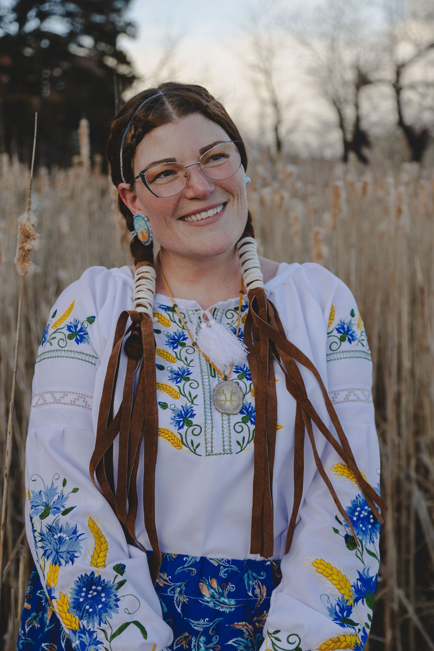 Smiling woman in embroidered Ukrainian vyshyvanka blouse with blue floral patterns, braids, and glasses outdoors.